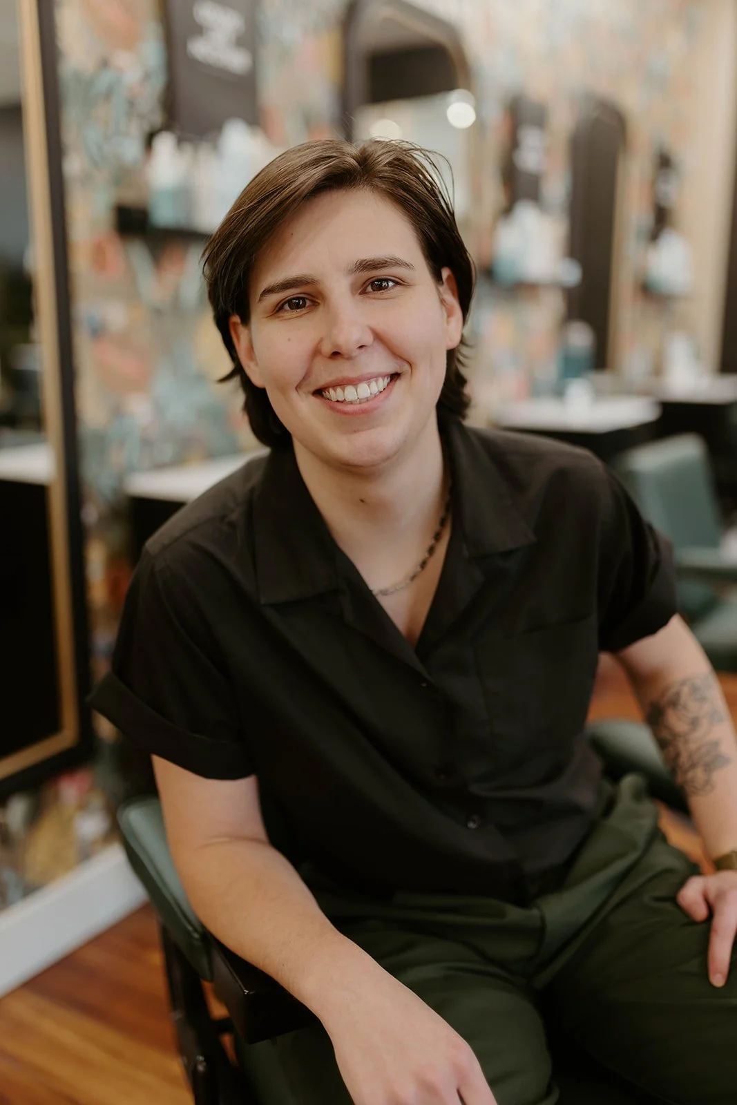 A young man with shoulder-length dark hair, smiling, wearing a black shirt, sitting in a barbershop or salon with shelves of hair products and taps in the background.