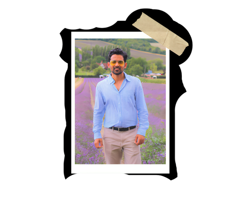 Man standing in a lavender field with green hills and houses in the background, wearing a blue striped shirt and beige pants, posed for a photo with a scenic landscape.