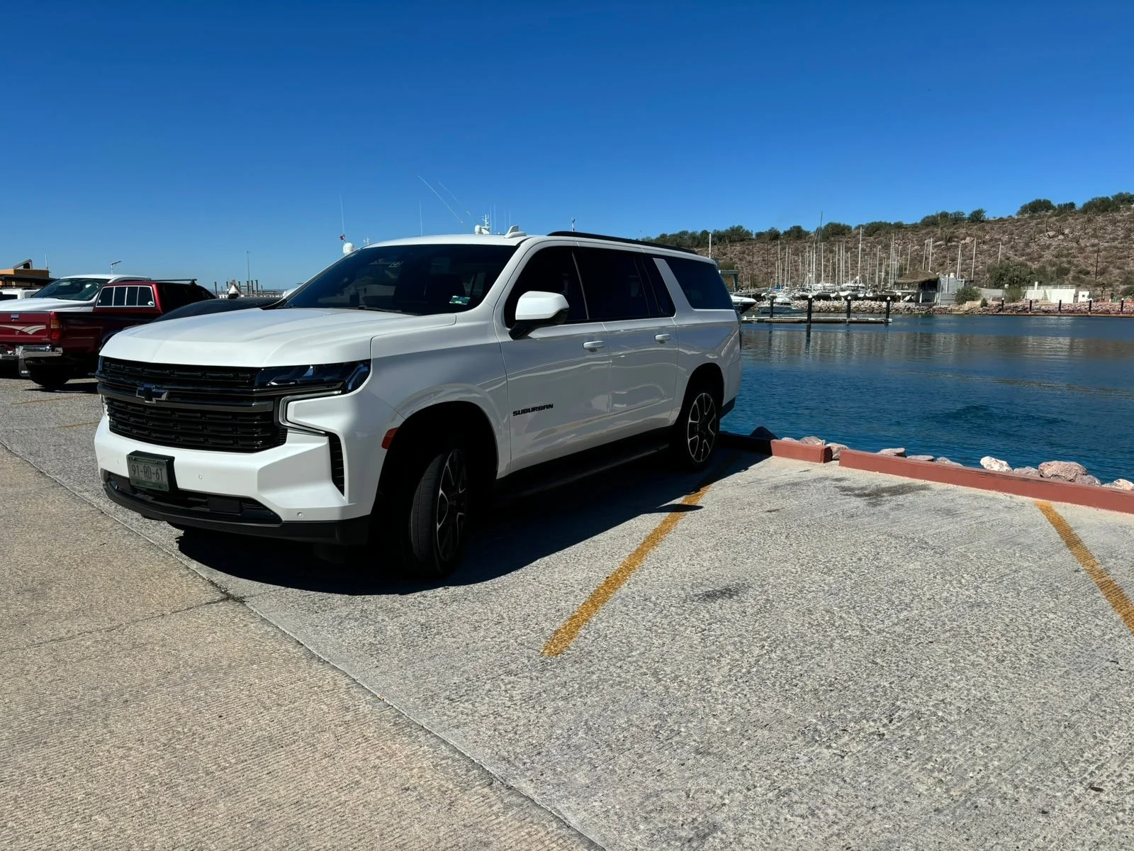 A white Chevrolet Suburban parked in a parking lot near a body of water with boats and a marina in the background.