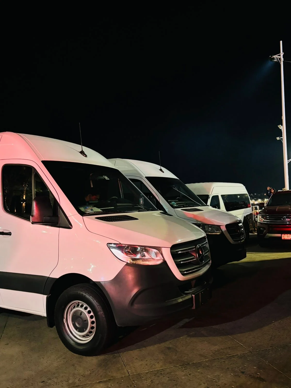 Nighttime parking lot with a row of vans and trucks, including a white Mercedes-Benz Sprinter in the foreground, illuminated by overhead lights, under a dark sky.