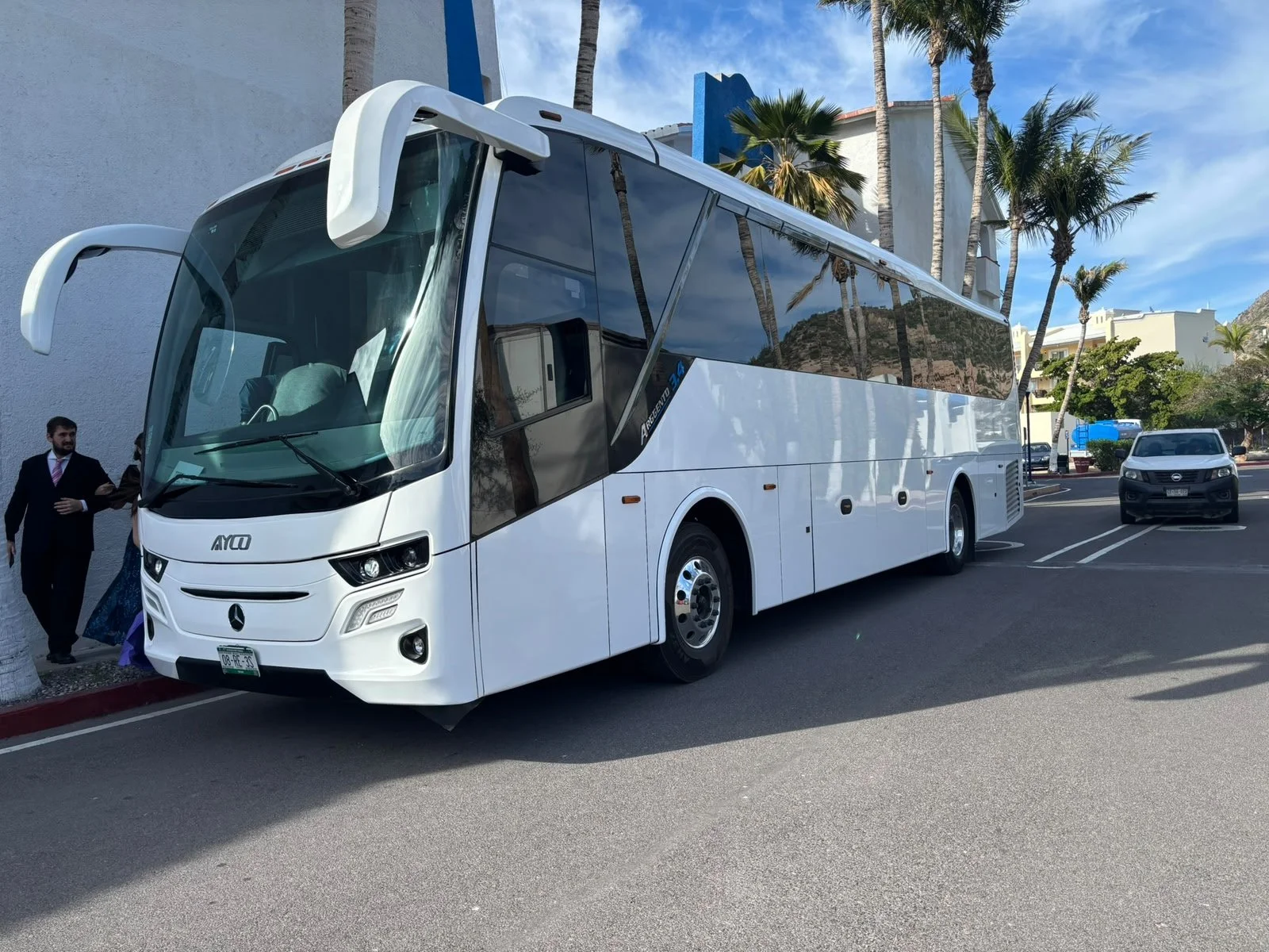 A white double-decker bus parked on the street near white buildings, palm trees, and a clear blue sky.