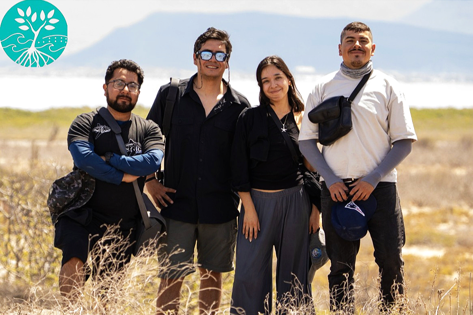 Four young adults standing outdoors in a grassy field with a cloudy sky in the background, smiling for the camera.