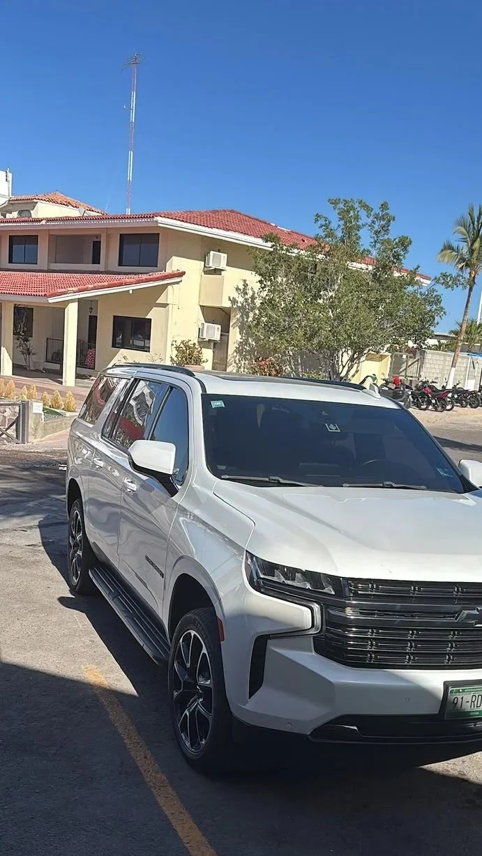 A white modern SUV parked in a parking lot with a beige residential building with red tile roof and a tree in the background under a clear blue sky.