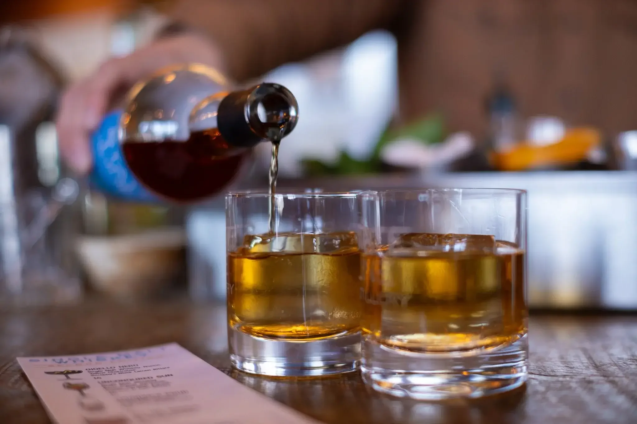 A person is pouring a dark liquor into a glass of ice, with a second glass of the liquor already filled on a wooden table in the foreground. A menu or receipt and a blurred background of a bar or restaurant setting are also visible.