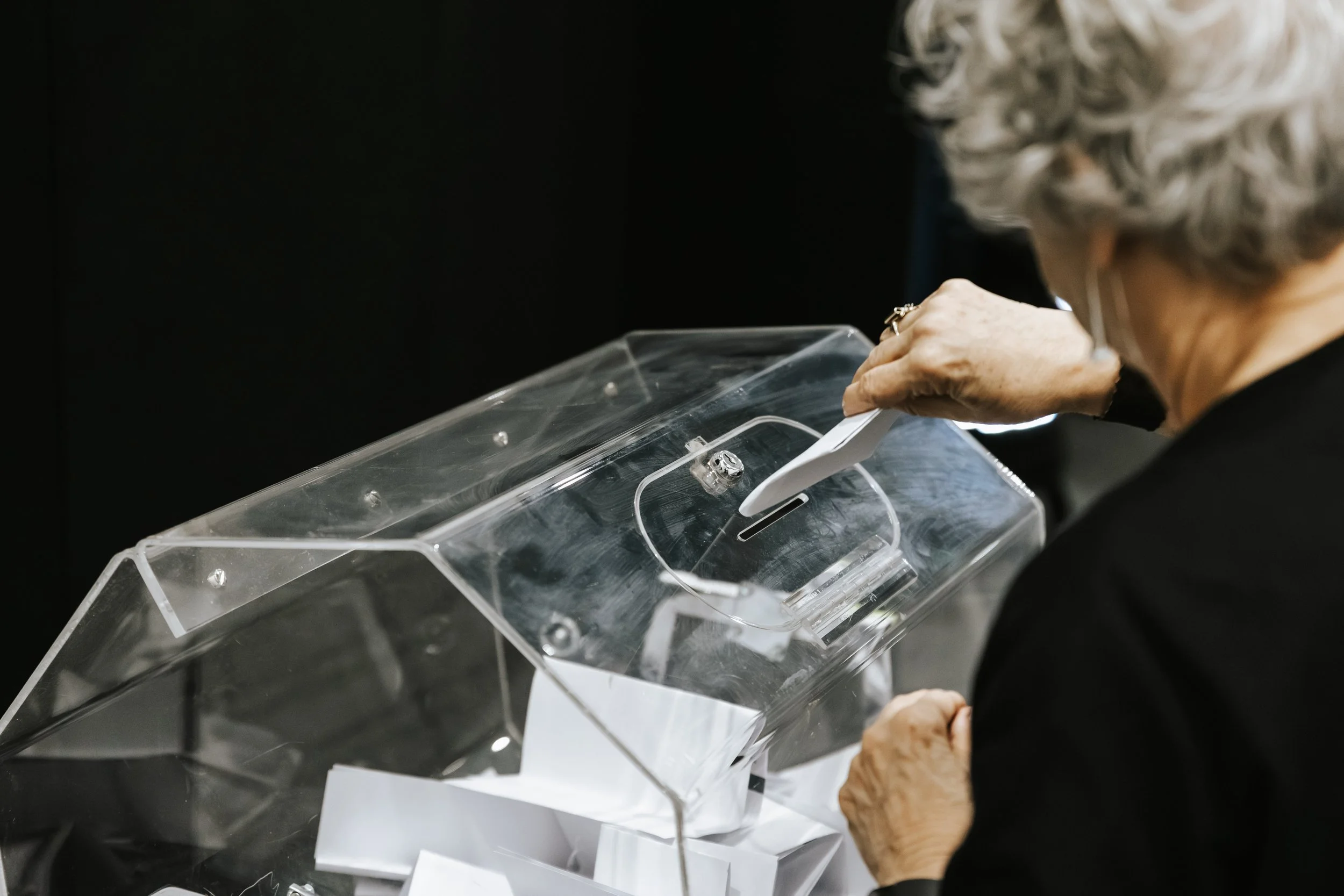 Two women sitting at a table, one with glasses and an embroidered jacket, talking and smiling, with papers and a small potted plant on the table.