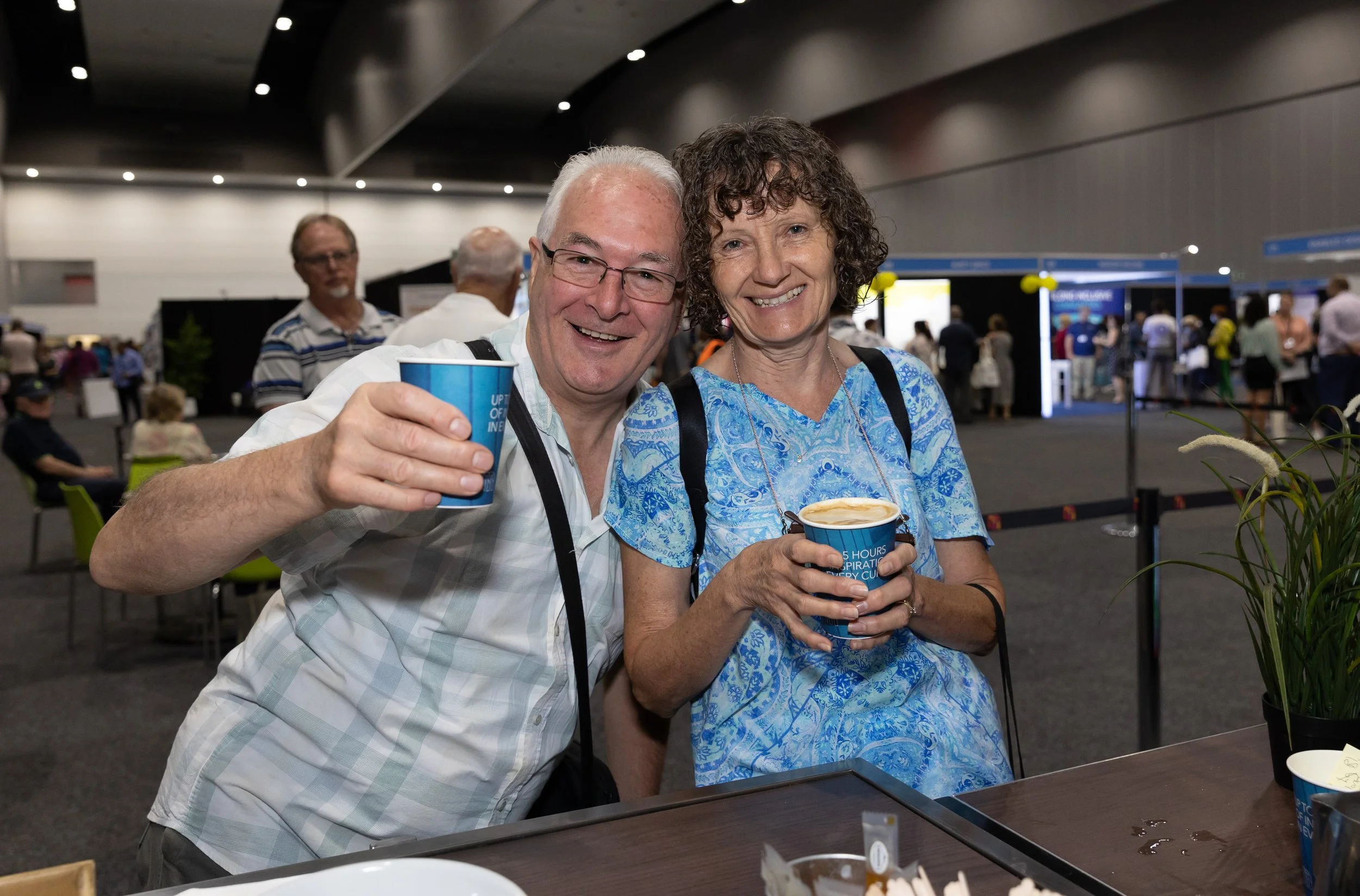 People seated and standing in a busy indoor café area with large orange signage on the wall reading 'Café Bar'. Some people are carrying bags, and there are signs indicating toilet facilities.