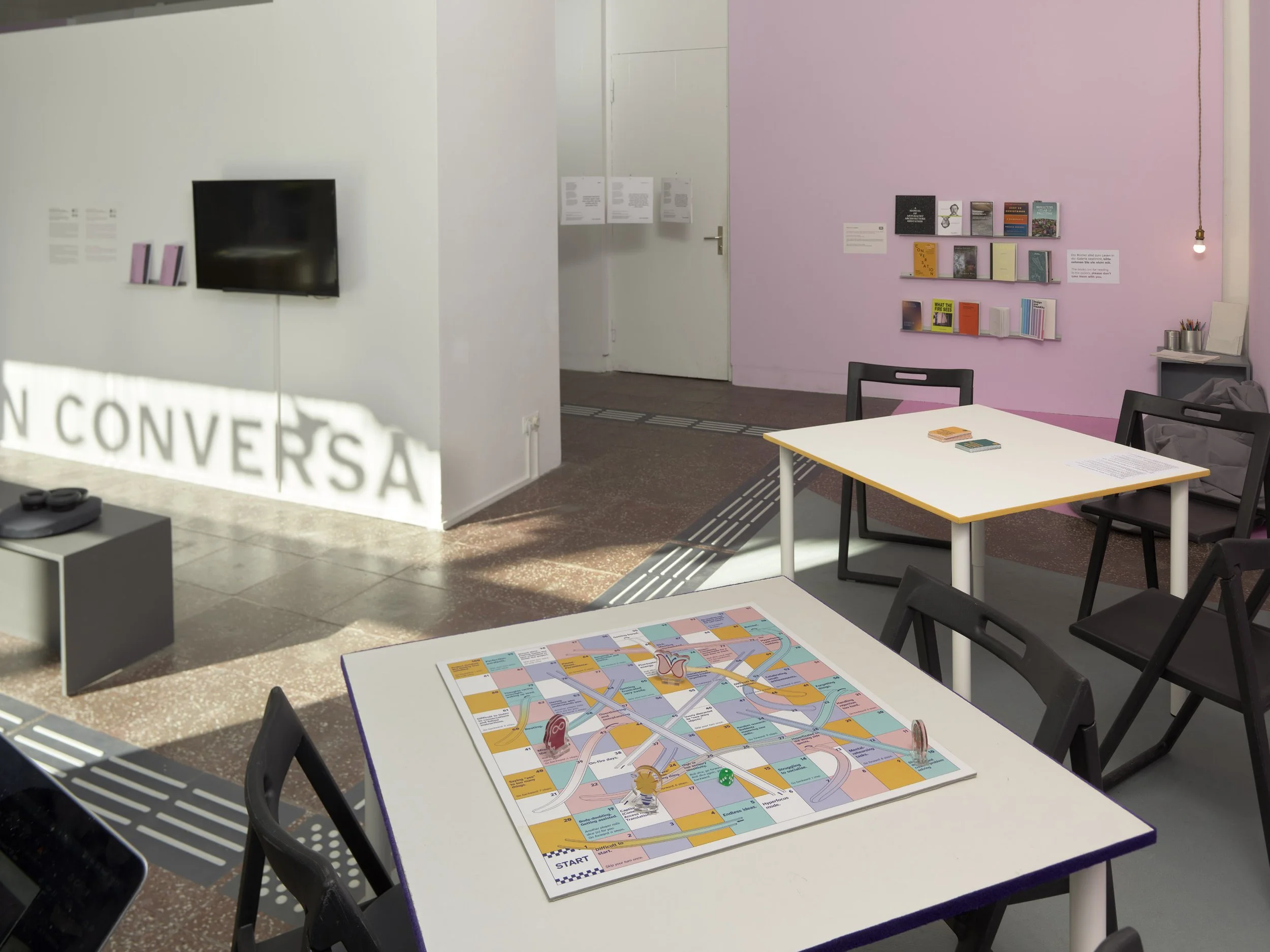 A wide shot of the table-top games, each one on a separate table. The image also features a monitor and printed transcription installation on the left and a reading corner on the far right corner. Photo: Eric Tschernow. Event: “In Conversation” exhib