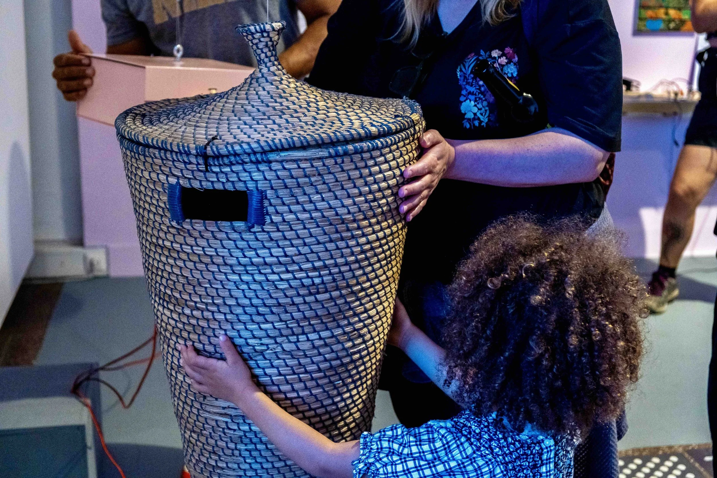 One child and two adults touch a hung rattan basket and a gift box. Photo: Mareike J. Lange. Event: “In Conversation” exhibition, 2025.