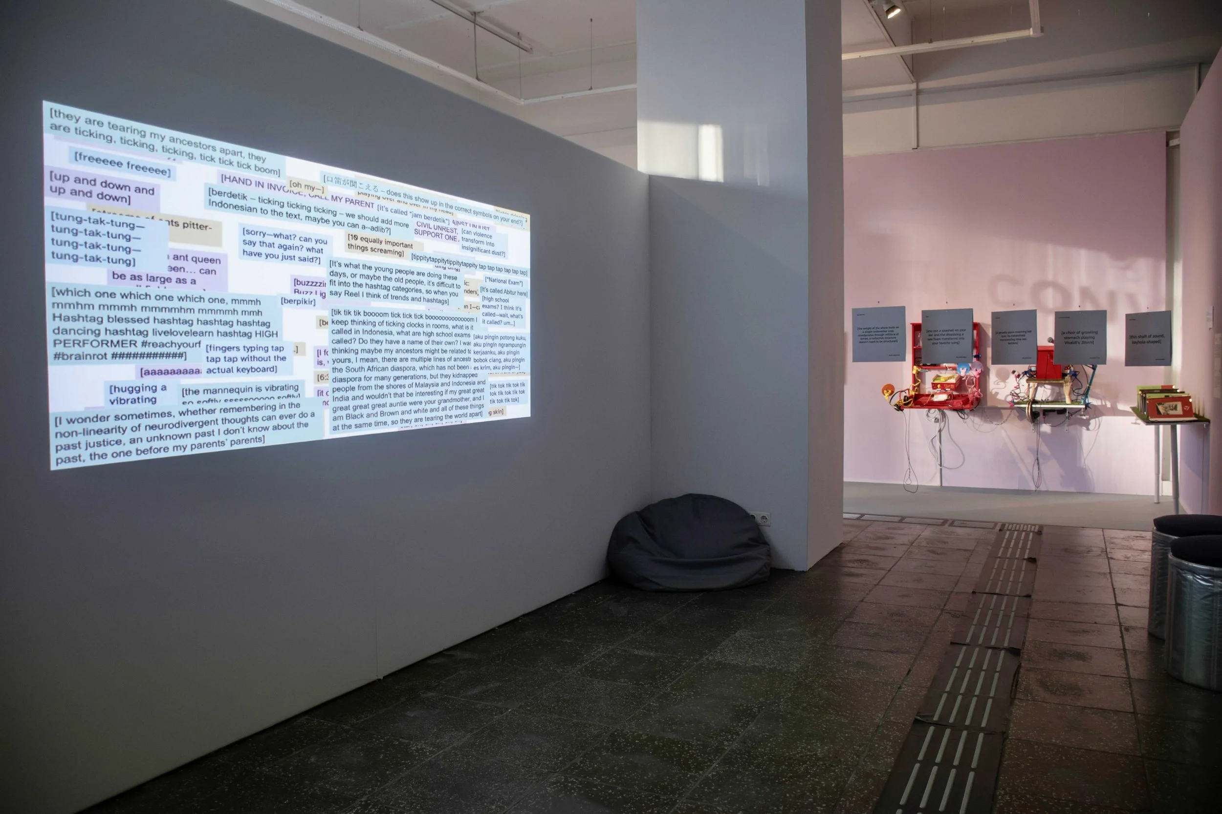 A visual projection on wall displaying a cloud of sentences, with bean bags and short stools around it. On the far right corner is installations of five hung vinyl covers, two sonic sculptures, and a zine. Photo: Dani Hasrouni: Event: “In Conversatio