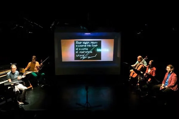 A front-angled camera shot. thingNY is on stage with captions on screen. The screen shows a film still that reads, “That night, there occurred the first of a series of mysterious crimes.” Captions, superimposed on the middle right, read, “intertitles