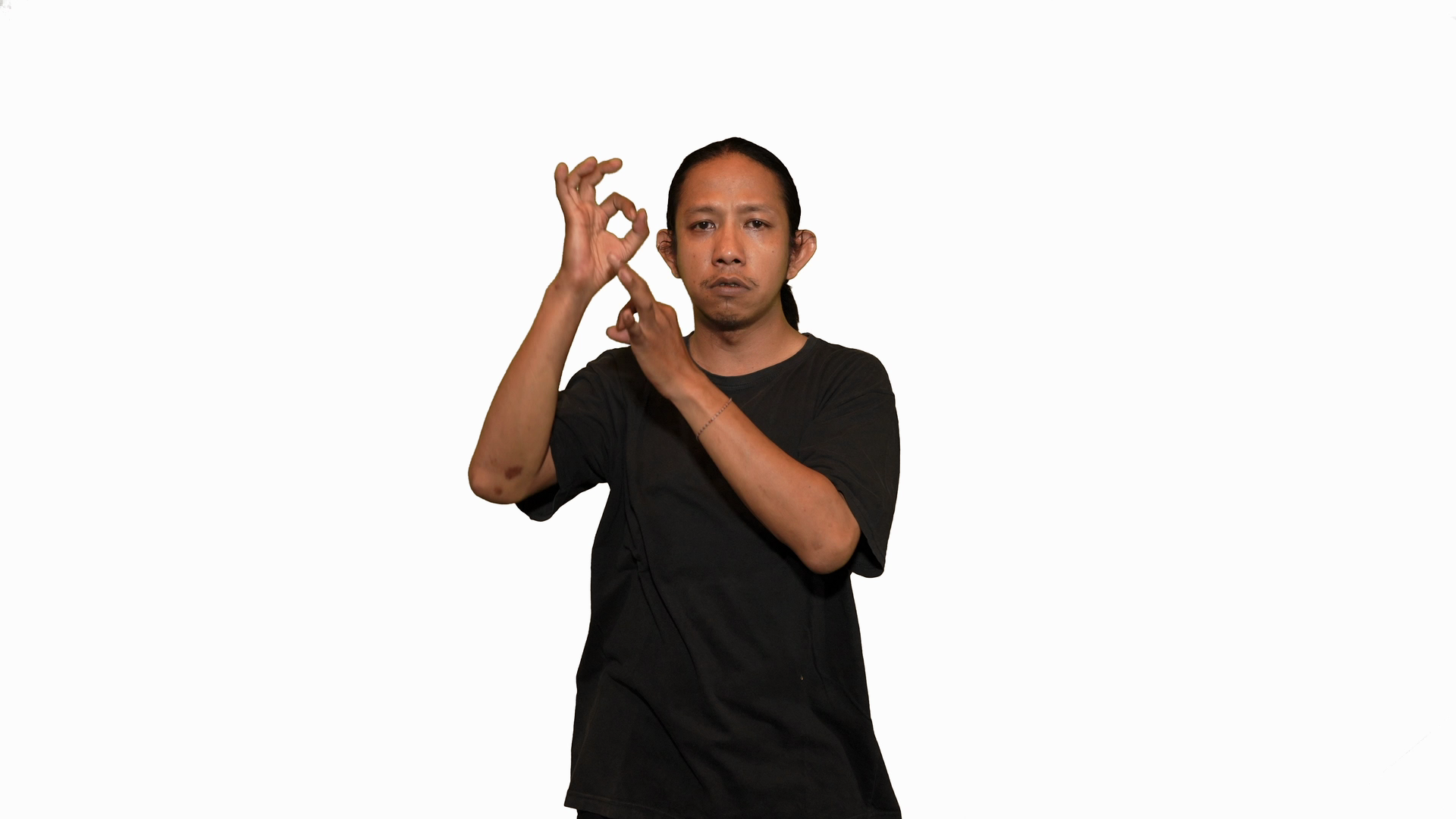 Snapshot from the film. Arief, a brown man with bunned hair wearing a black shirt, poses his hands on the right side of his head. His hands display open palms with the index and thumb meeting.