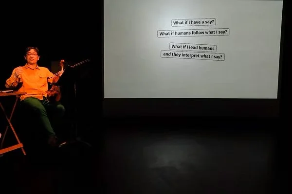 A front-angled camera shot. thingNY is on stage with captions on screen. The screen shows captions that read, “What if I have a say? What if humans follow what I say? What if I lead humans and they interpret what I say?” On the screen’s left side is