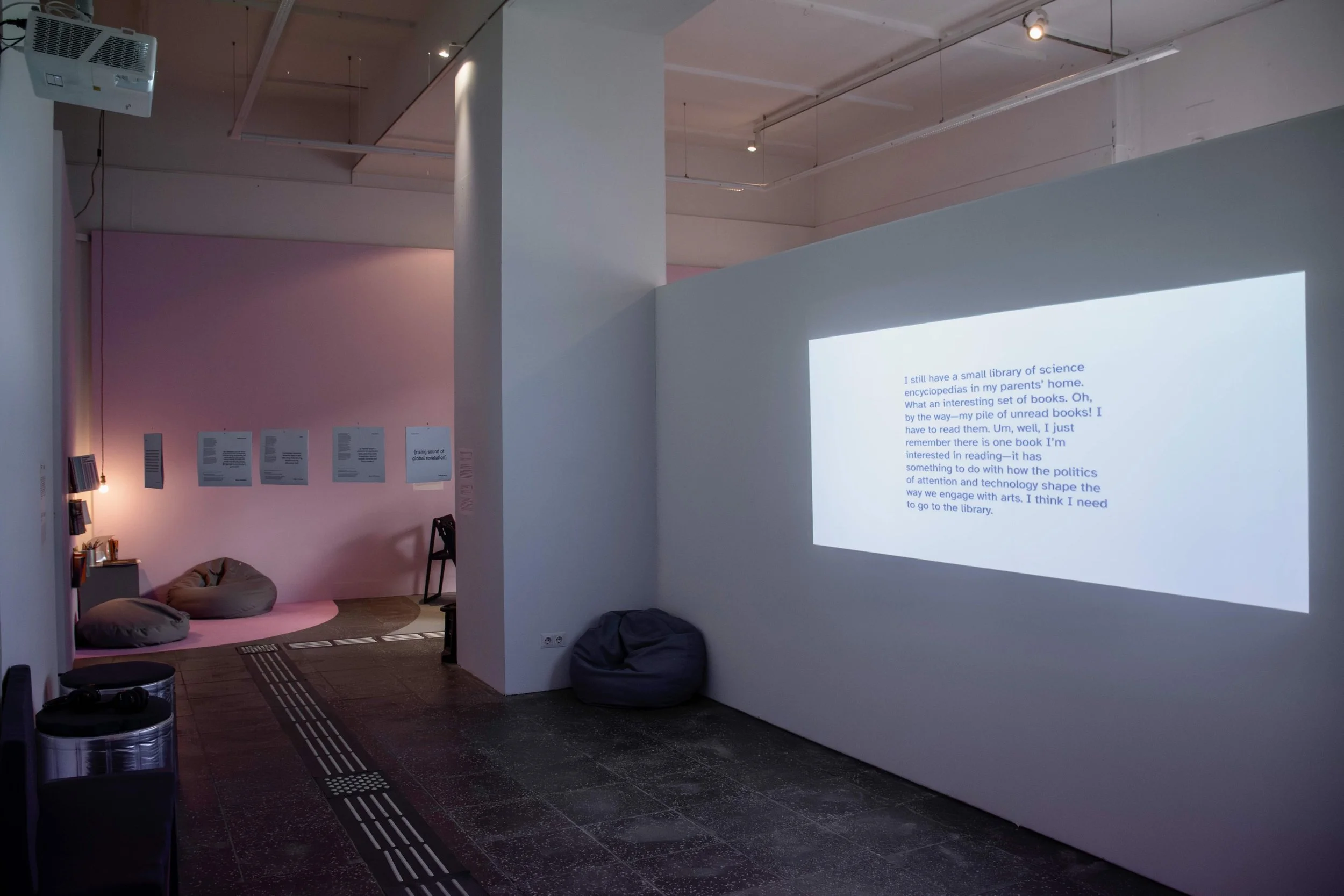 A visual projection on a wall displaying a text paragraph, with bean bags and short stools around it. On the far left corner is an installation of five vinyl covers and a reading corner. Photo: Dani Hasrouni: Event: “In Conversation” exhibition, 2025