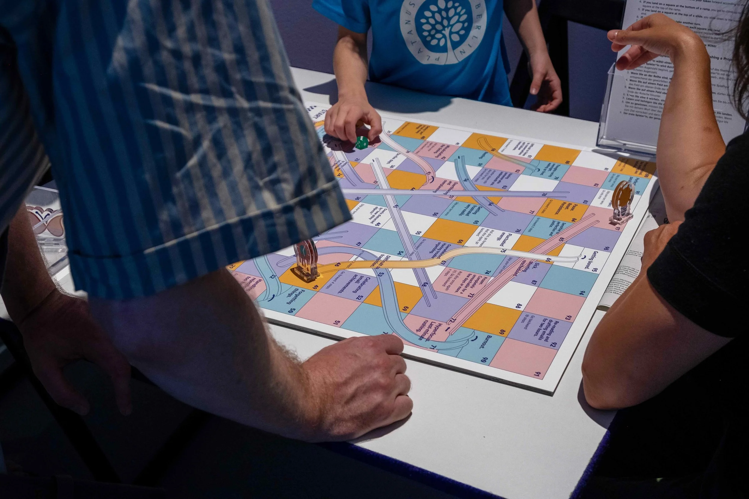 Two adults and one kid play the board game. Photo: Mareike J. Lange. Event: “In Conversation” exhibition, 2025.