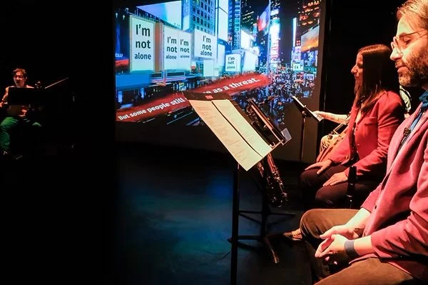 A right-angled camera shot. thingNY is on stage with captions on screen. The screen shows many buildings and digital billboards that read, “I’m not alone” and a street covered with captions, “But some people still see me as a threat.” On the screen’s