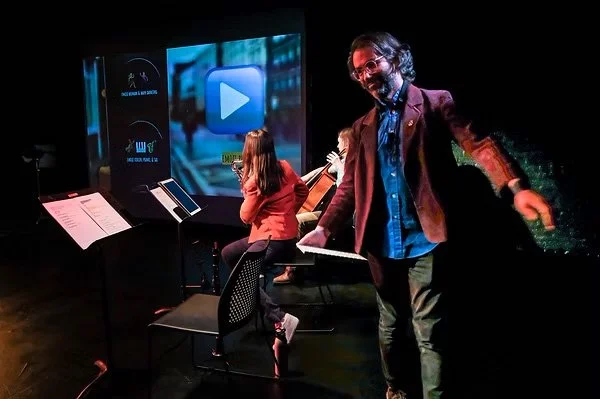 A right-angled camera shot. thingNY is on stage with captions on screen. On the screen’s right side, front to back, Dave Ruder stands with both arms half-raised while holding a wireless keyboard, Erin Rogers plays a saxophone while facing the screen,