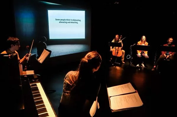 A left-angled camera shot. thingNY is on stage with captions on screen. The screen shows a male sitting and looking down—in front of him is a white screen with captions that read, “Some people think I’m distracting, alienating and detaching” ended wi