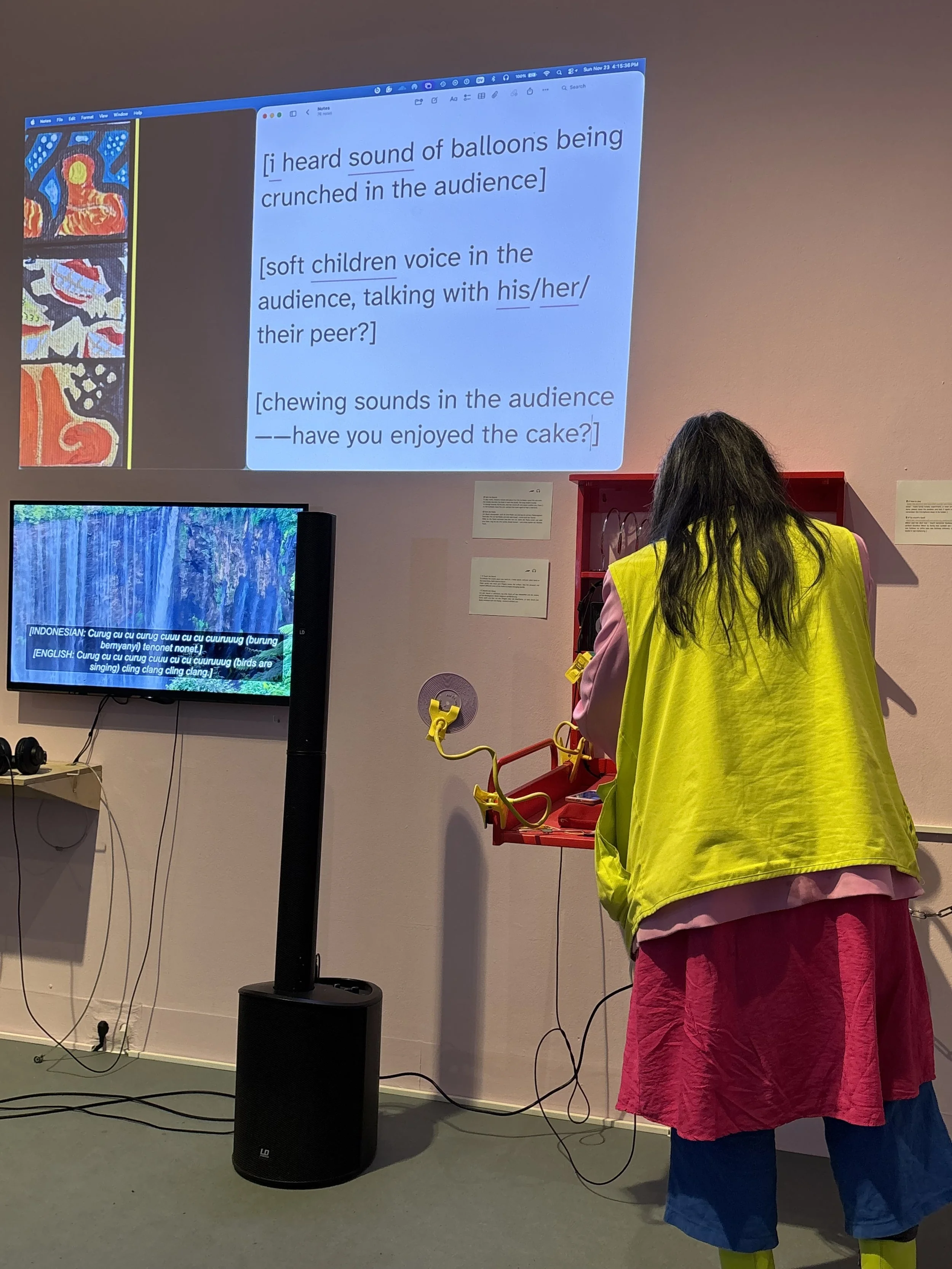 Sound captions projected on a wall. They interpreted the overall soundscape. They read, “[i heard sound of balloons being crunched in the audience] [soft children voice in the audience, talking with his/her/their peer?] [chewing sounds in the audienc