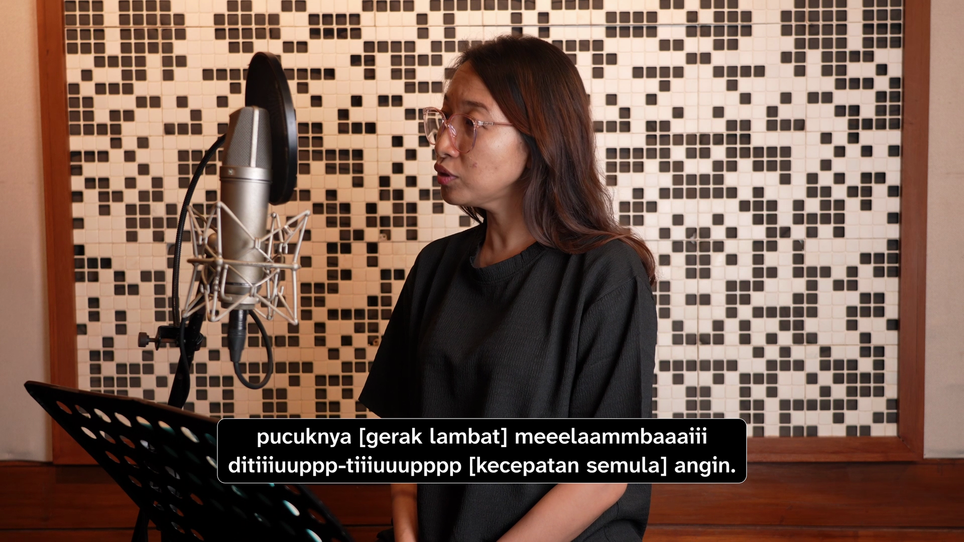Snapshot from the film displaying Bawien, a brown woman with shoulder-length red hair wearing a black shirt, singing and sitting in front of a microphone. The sound captions read, “pucuknya [gerak lambat] meeelaammbaaaiii ditiiiuuppp-tiiiuuupppp [kec