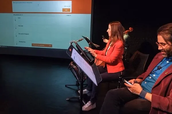 A right-angled camera shot. thingNY is on stage with captions on screen. The screen shows a list of captions generated by the audience. On the screen’s right side, front to back, Dave Ruder types on his phone, Erin Rogers types on a wireless keyboard