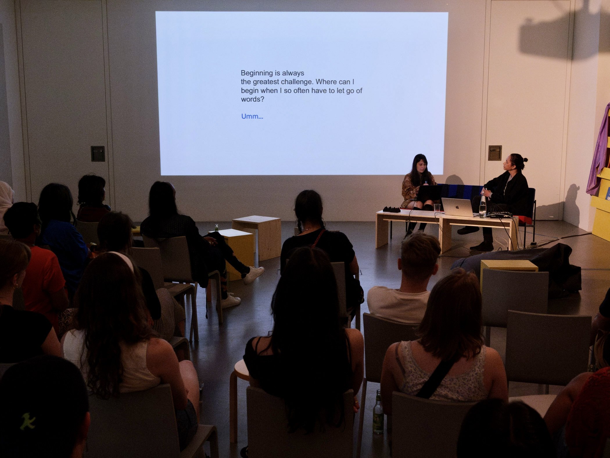 Simoné and Jay on stage performing a projected text while sitting on a colorful couch. The audience sits dispersely, watching. Photo: Eunice Maurice / DAAD Artists-in-Berlin.