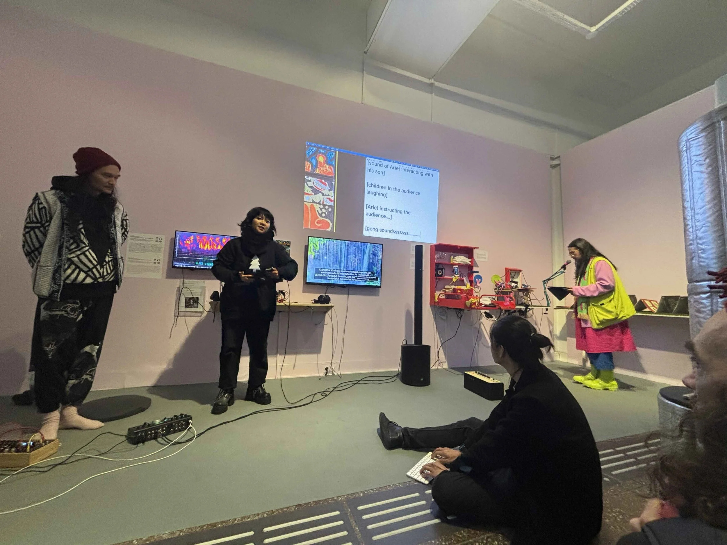 Sound captions projected on a wall. They interpreted sounds performed by Ariel, Dea, and Morgan, and the overall soundscape. They read, “[sound of Ariel interacting with his son] [children in the audience laughing] [Ariel instructing the audience....