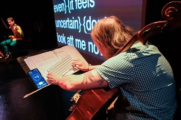 A right-angled camera shot. thingNY is on stage with captions on screen. On the screen’s left side is a blurred shot of Jeffrey Young playing violin. On the screen’s right side is a focused shot of Andrew Livingston typing a keyboard on a music stand