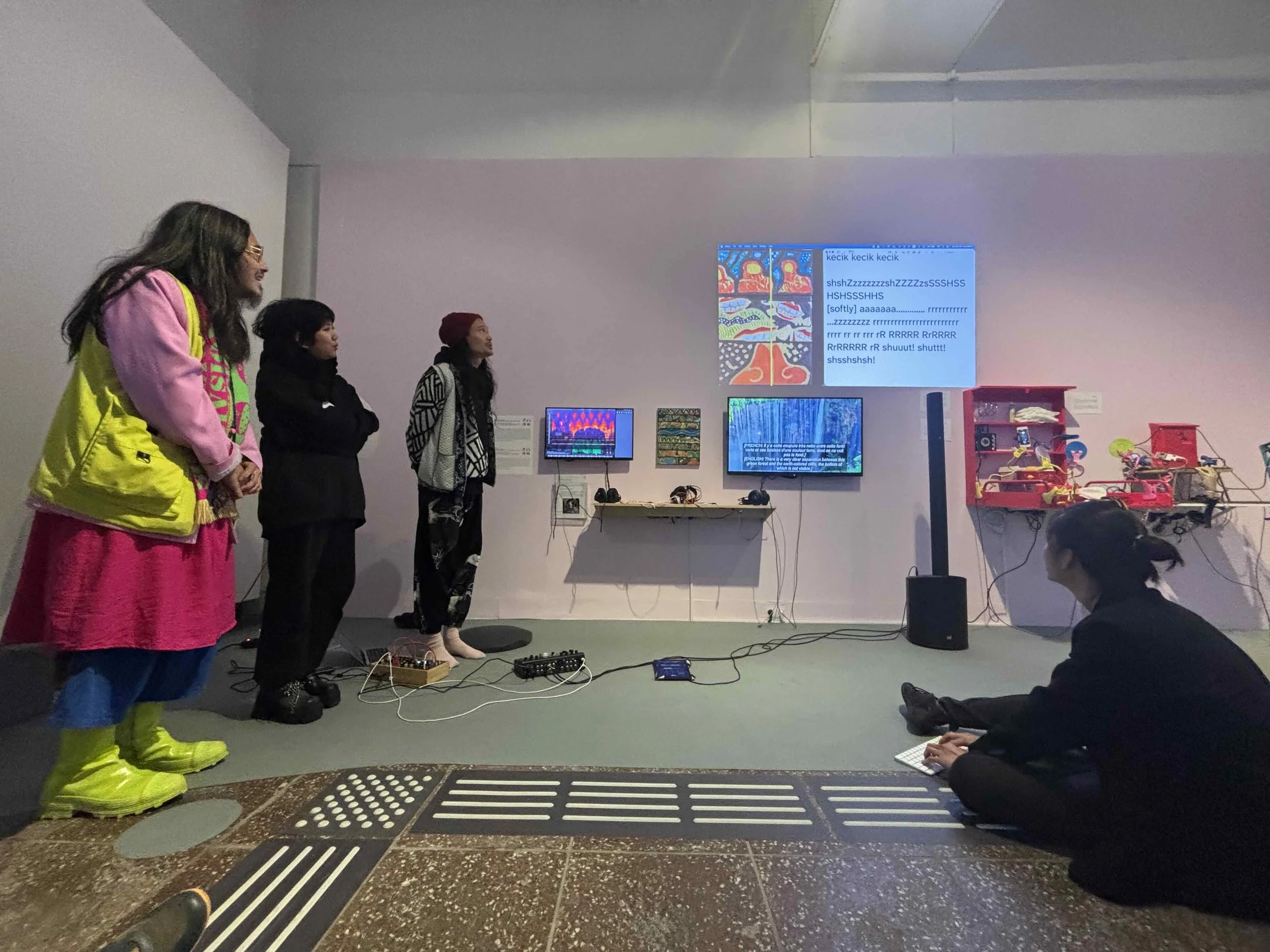 Sound captions projected on a wall. They interpreted three human voices performed by Ariel, Dea, and Morgan. They read, “kecik kecik kecik shshZzzzzzzzzshZZZZzsSSSHSSHSHSSHHS [softly] aaaaaa.............. rrrrrrrrrrrrr ...zzzzzzzzz rrrrrrrrrrrrrrrrrr