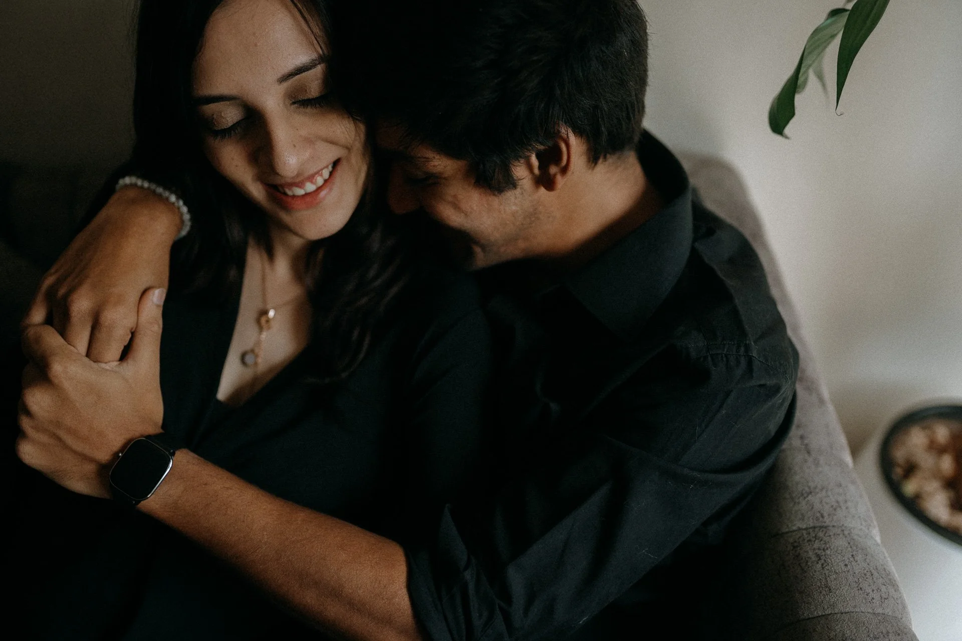 A smiling woman with dark hair and a man with dark hair hugging on a couch, sharing a joyful moment.
