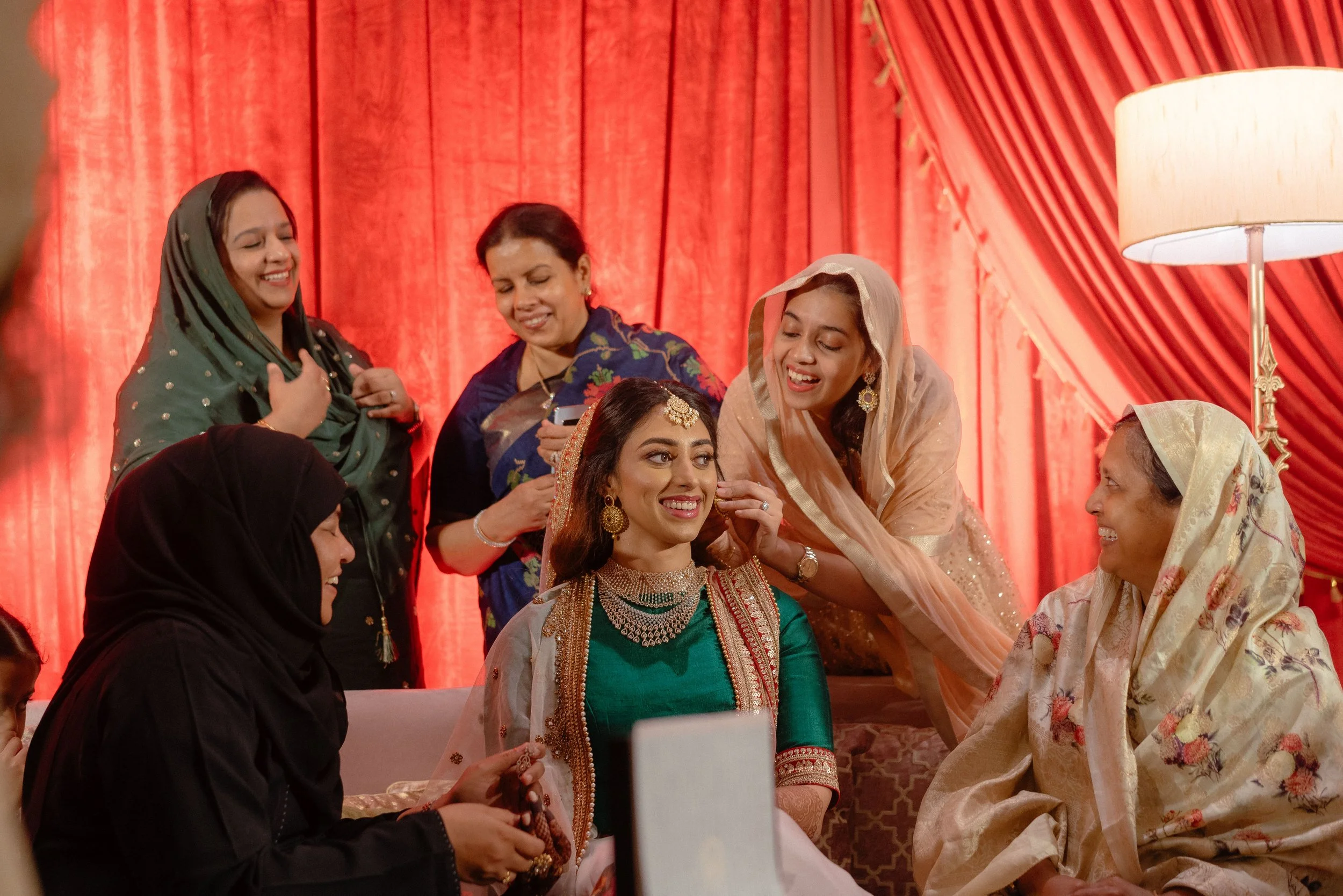 Group of women in traditional Indian attire at a wedding, with one woman seated in the center being groomed, surrounded by friends and family in vibrant clothing, in front of red curtains.