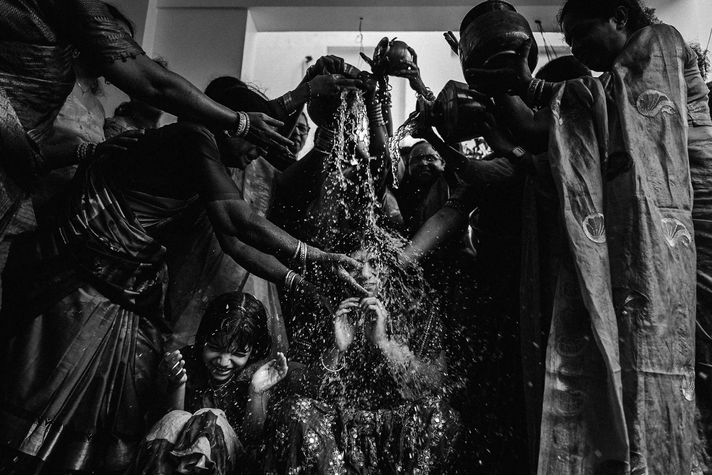 People participating in a religious or cultural ritual involving pouring water over a woman, with others helping and observing, in a dimly lit indoor setting.