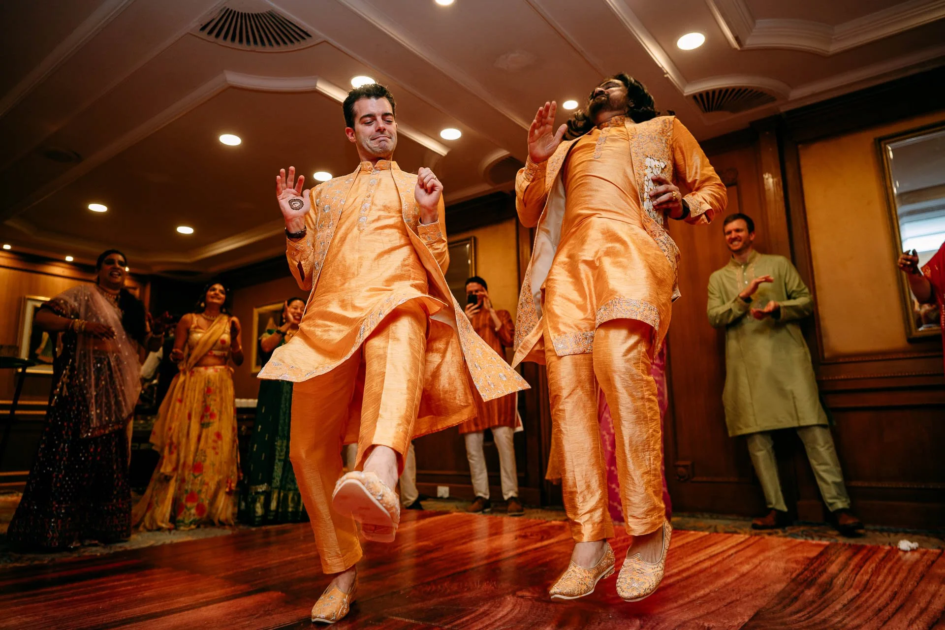 Two men dressed in gold Indian traditional attire dancing at a wedding celebration, with guests clapping and smiling in the background.