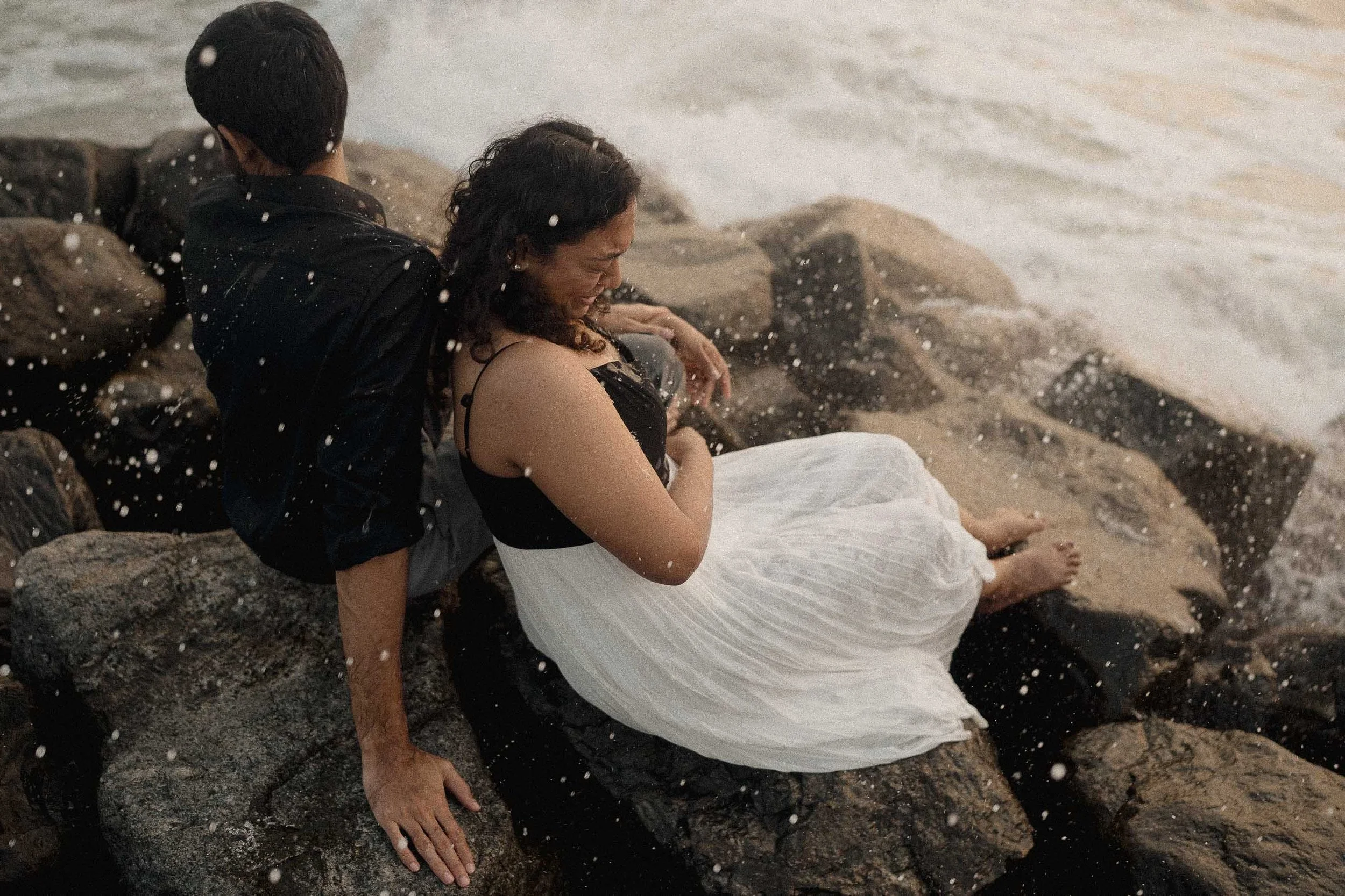 A man and woman sitting on rocks by the water. The woman is crying and the man is supporting her, with a splash of water around them.