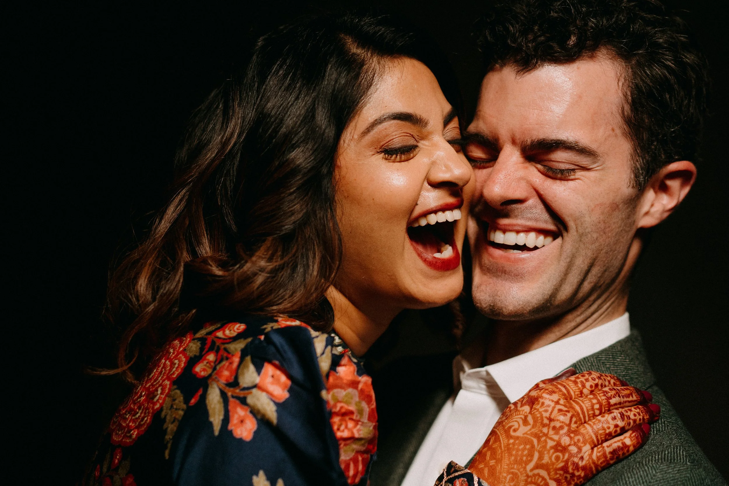 A man and woman laughing and smiling closely, with the woman wearing a floral dress and the man in a suit, against a black background.