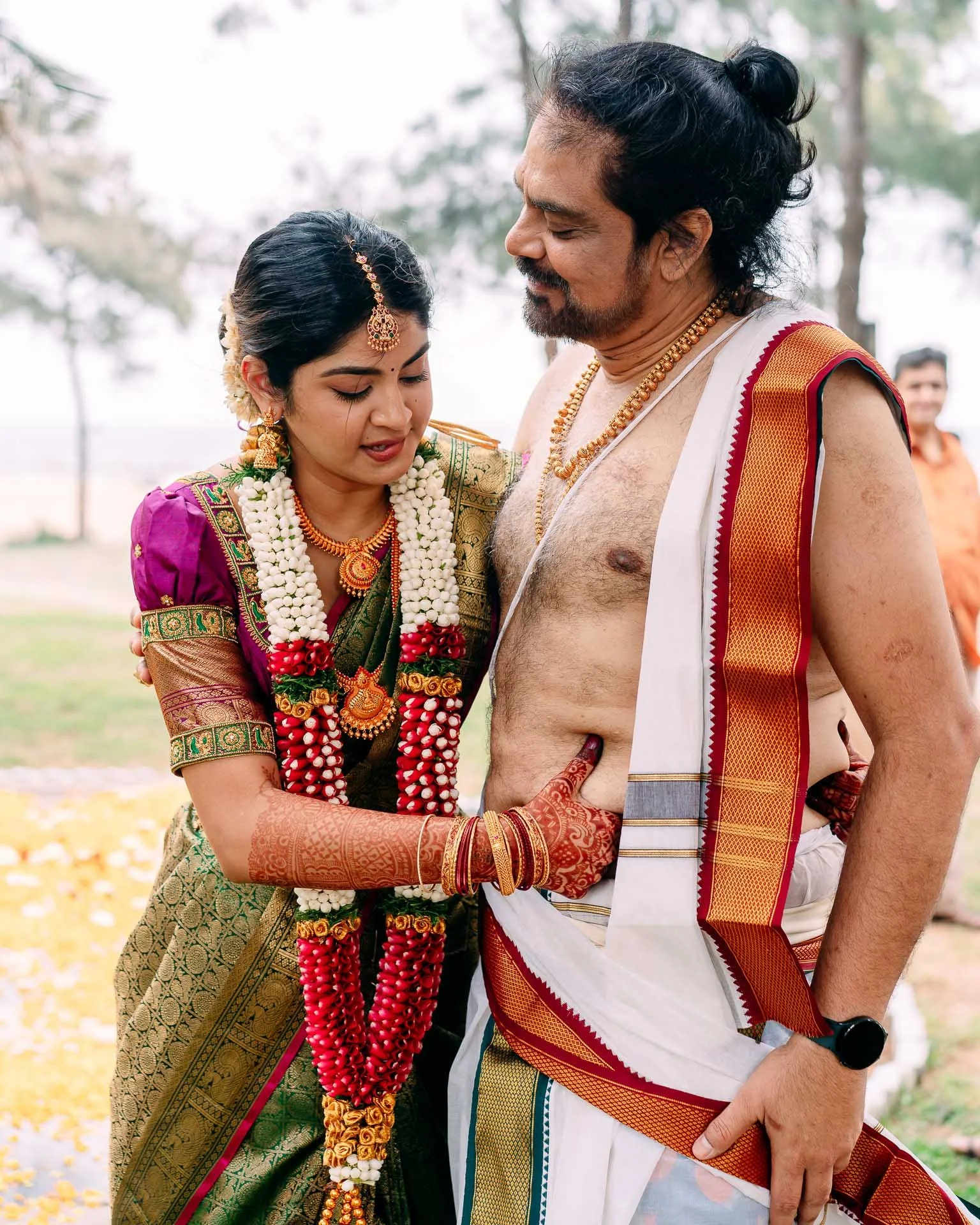 A couple dressed in traditional South Indian wedding attire participating in a wedding ritual outdoors. The bride is wearing a purple and gold saree with gold jewelry and floral garlands, and the groom is wearing a white dhoti with a sleeveless shawl