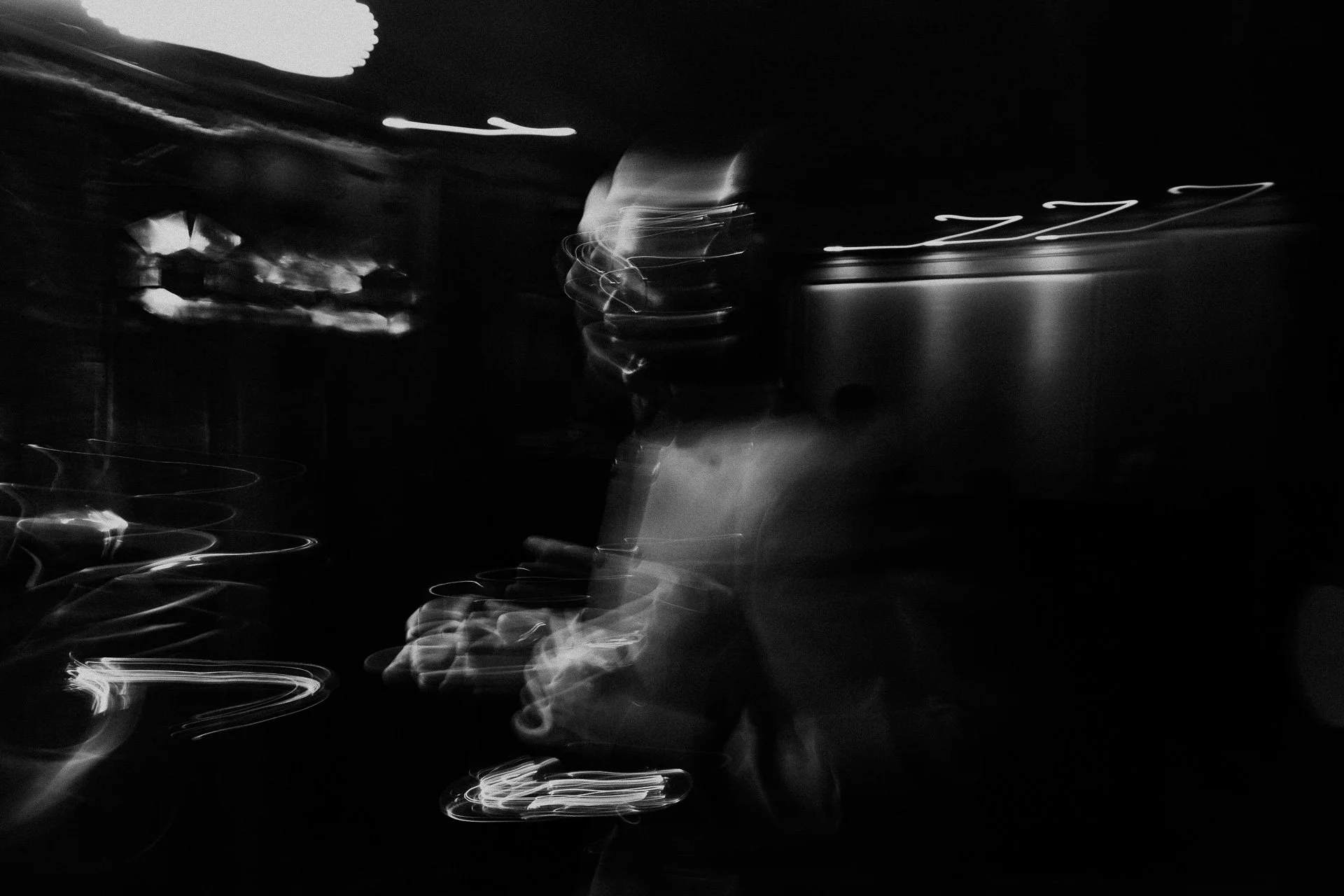 Blurred black and white photo of a person wearing glasses in a dark environment, with streaks of light creating abstract patterns.