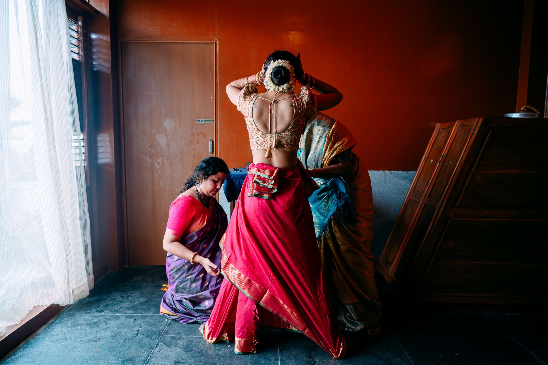 Women in traditional Indian attire preparing for a wedding or cultural ceremony indoors near a window and wooden wall.