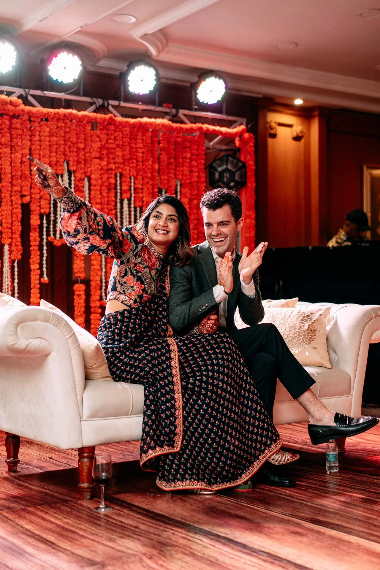 A man and woman sitting on a cream-colored sofa, smiling and celebrating at a festive event with orange marigold flower decorations and bright lights behind them.