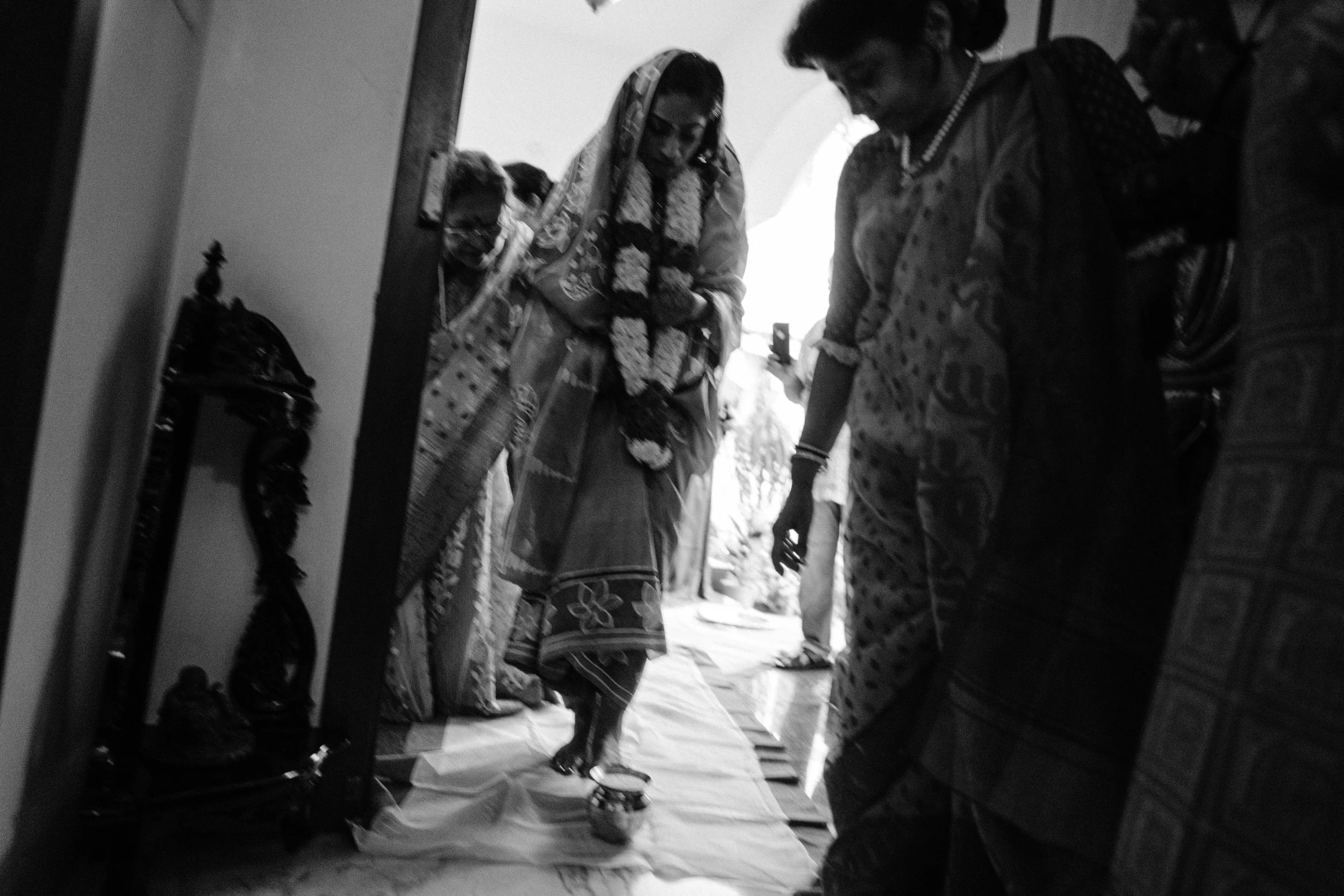 Group of women in traditional Indian attire gathered inside a house, with one woman balancing on a small platform, possibly participating in a religious or cultural ritual.