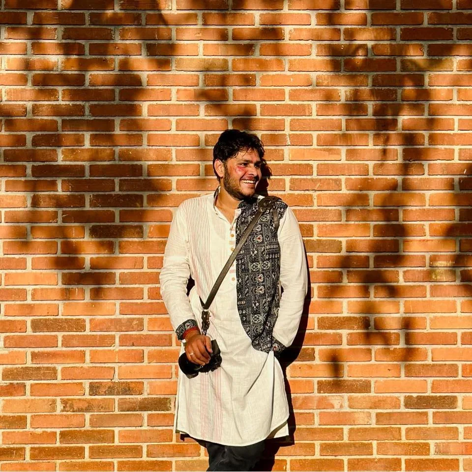 Man standing against a brick wall with shadows, wearing traditional Indian attire and holding a camera