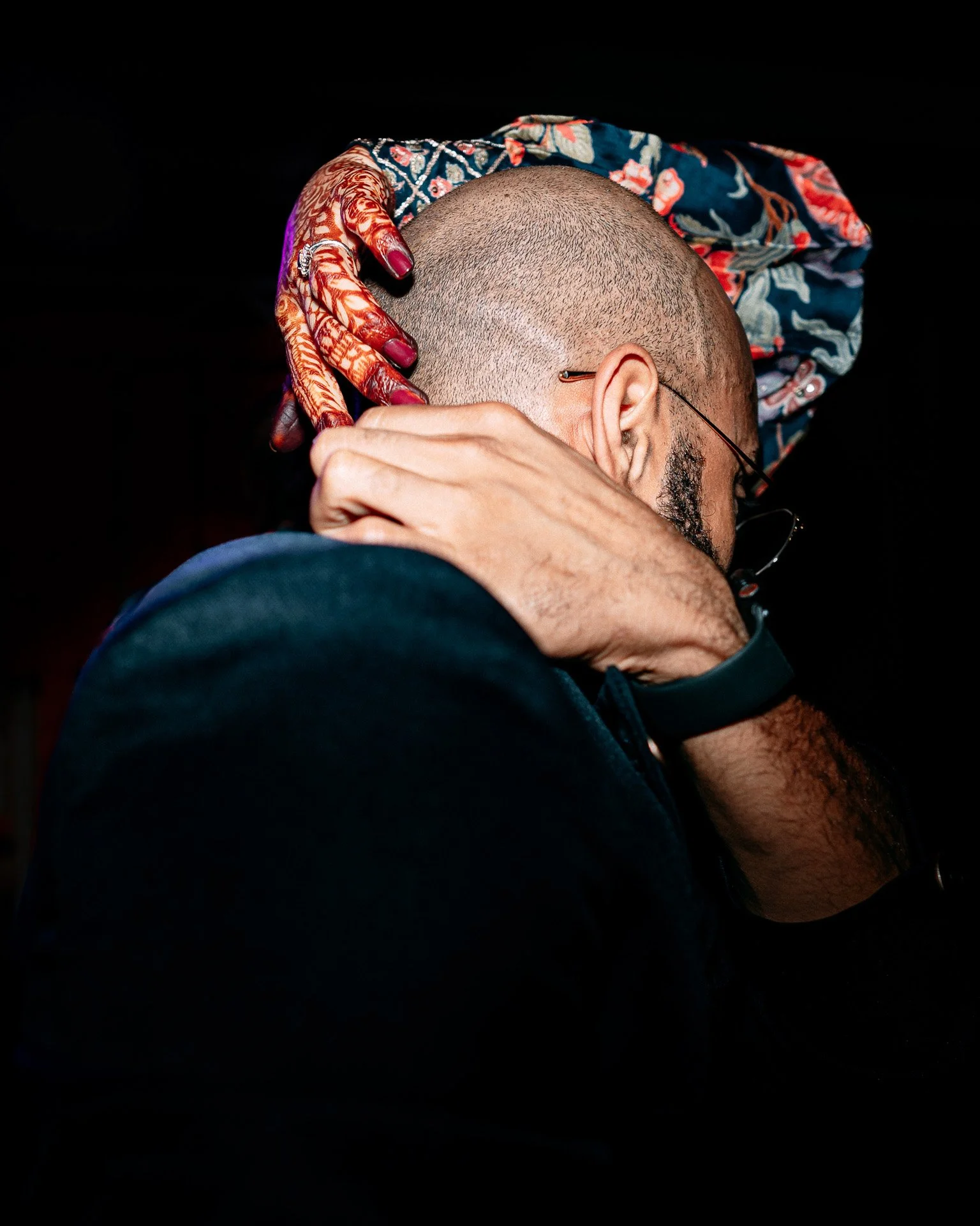 A person holding another person's head with their hand, both in an embrace. One has a decorative shawl, and the other's head is shaved. The scene is intimate and emotional.