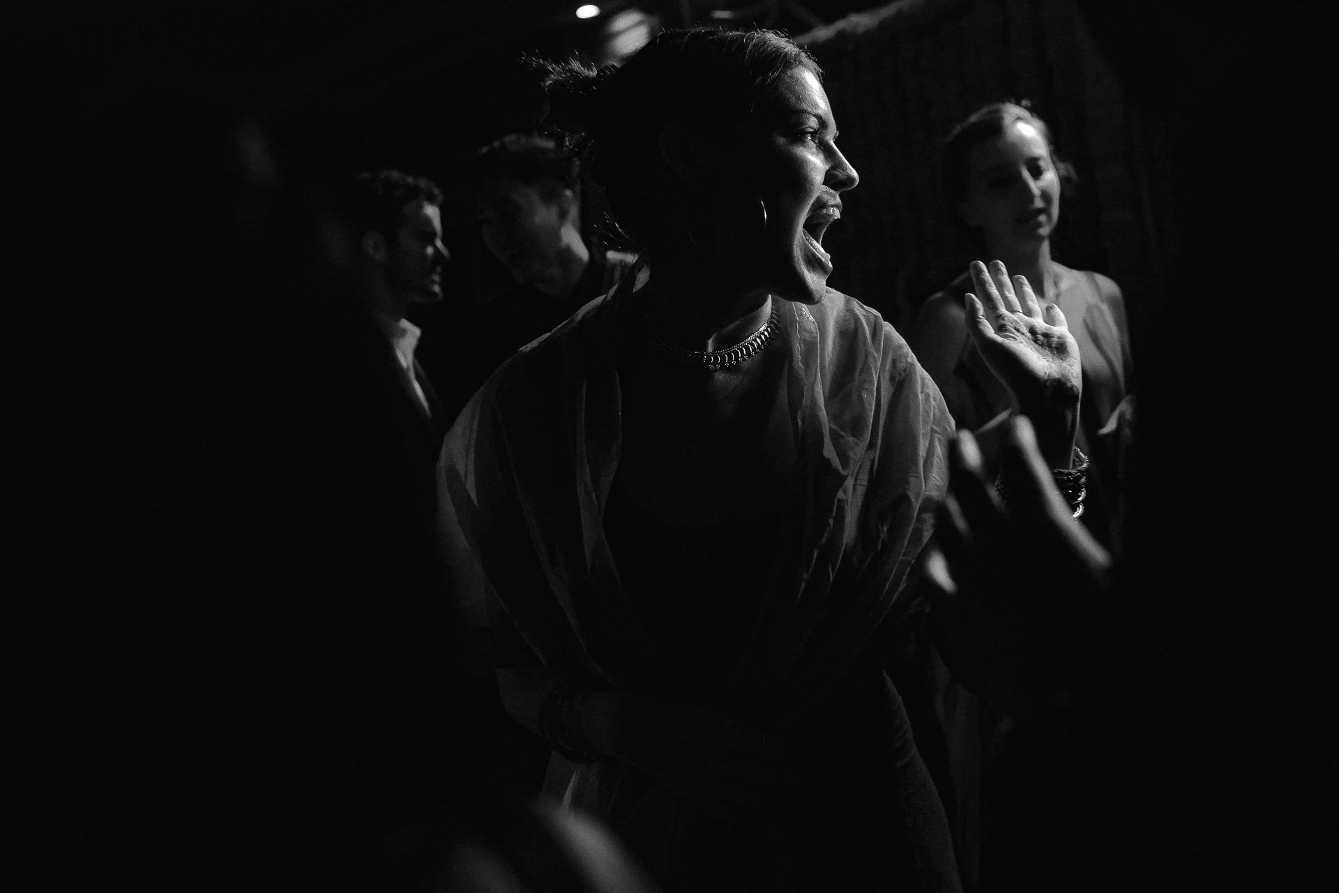 A woman with a short hairstyle and jewelry is laughing or talking animatedly, with other people in the background, in black and white.