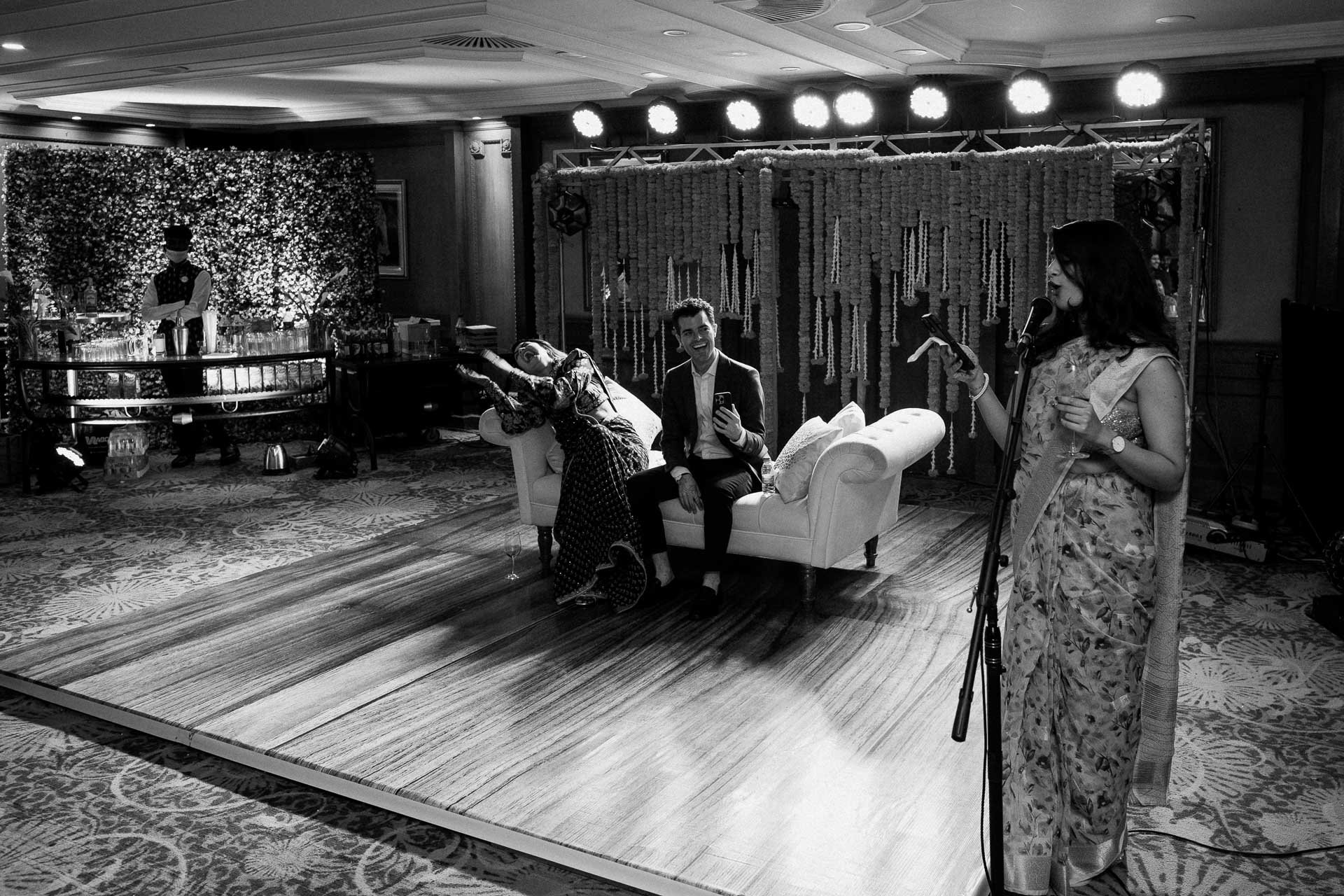 Black and white photograph of a wedding reception in a ballroom with a decorated stage. A woman in sari is speaking at a microphone, a bride and groom sit on a couch, smiling, with the groom holding a phone. A bartender is at the bar in the backgroun