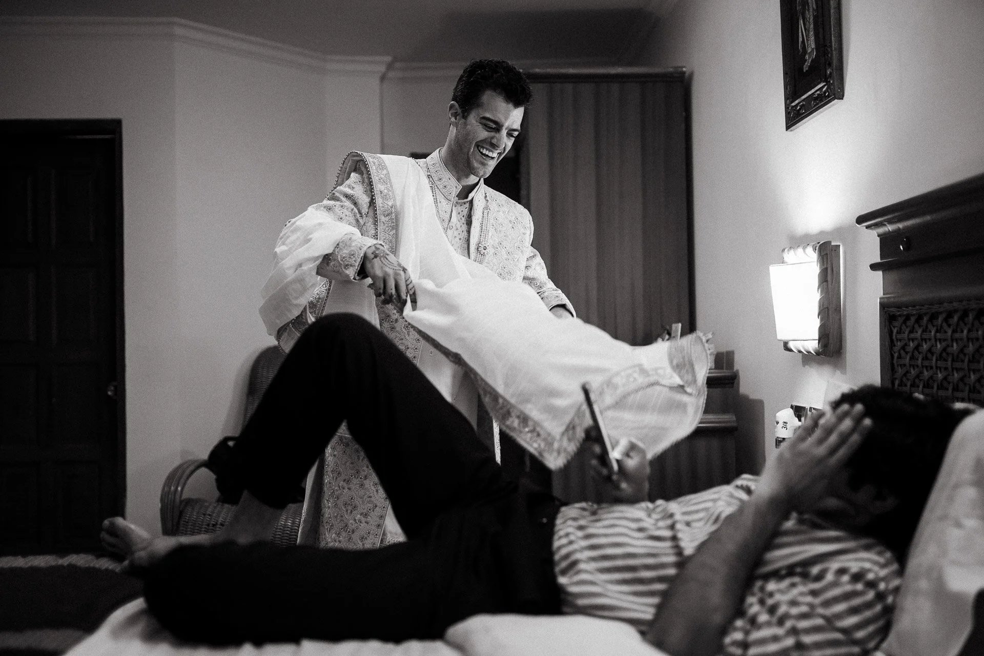 A man lying on a bed with a striped shirt and a cap, writing or drawing on a notepad while a woman dressed in traditional South Asian attire, called a saree, stands beside the bed smiling and pulling the saree. The scene is in a bedroom with wooden f
