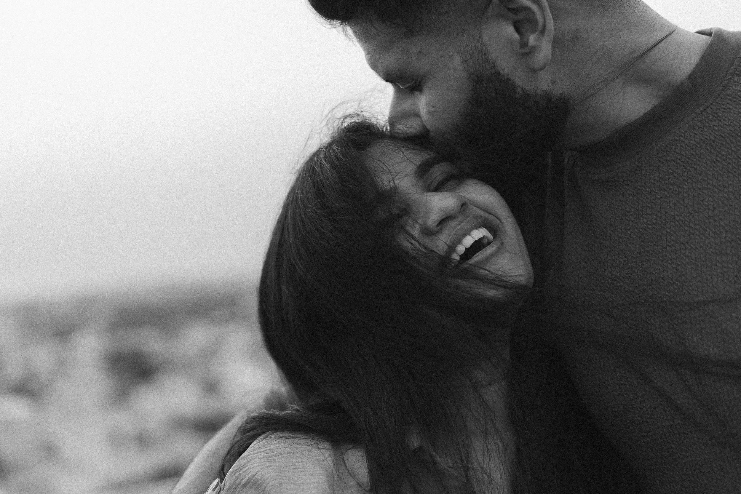A black-and-white photo of a joyful woman being embraced and kissed on the forehead by a man, both smiling and showing affection.