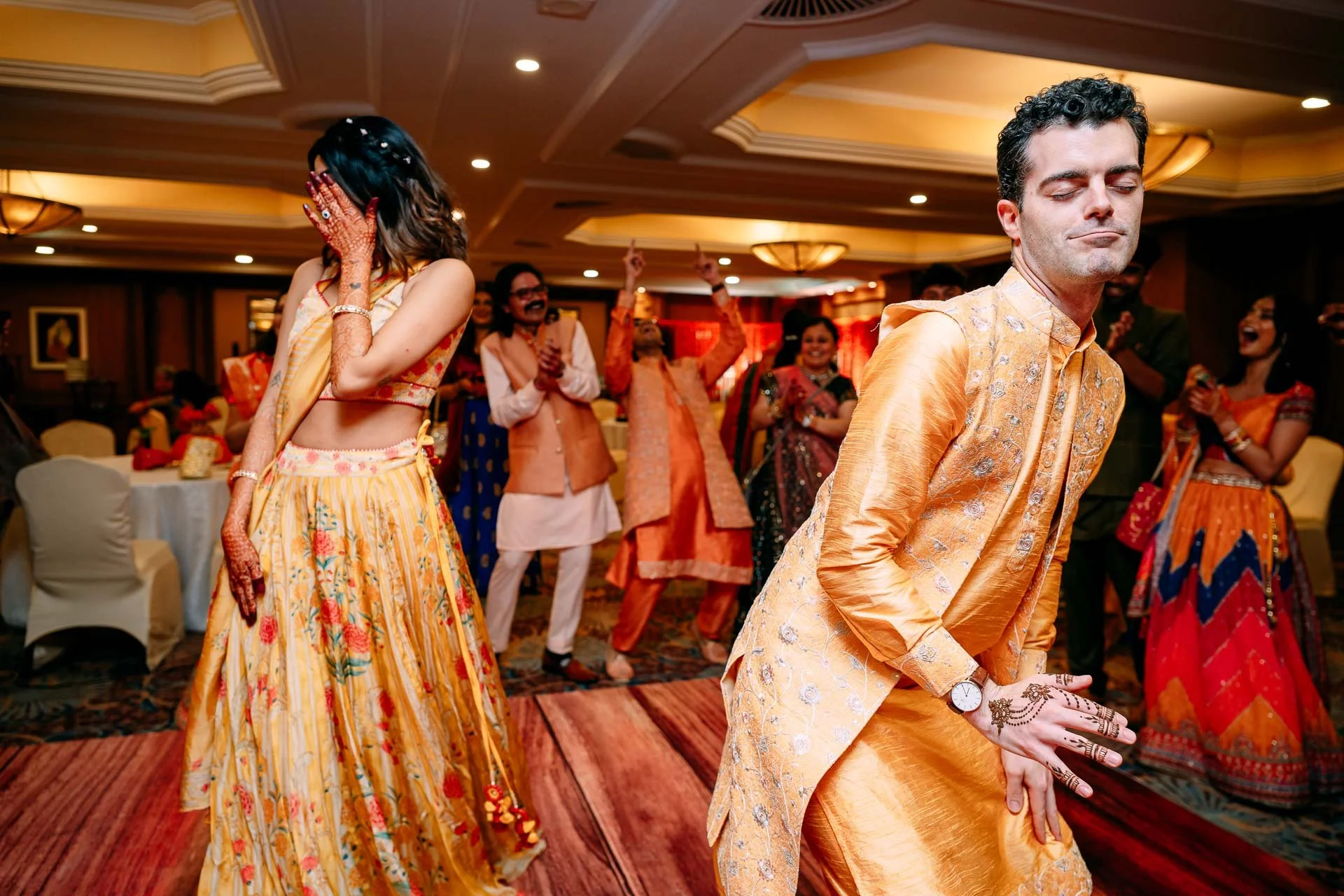 People dancing and celebrating at an indoor Indian wedding reception, wearing traditional colorful attire.