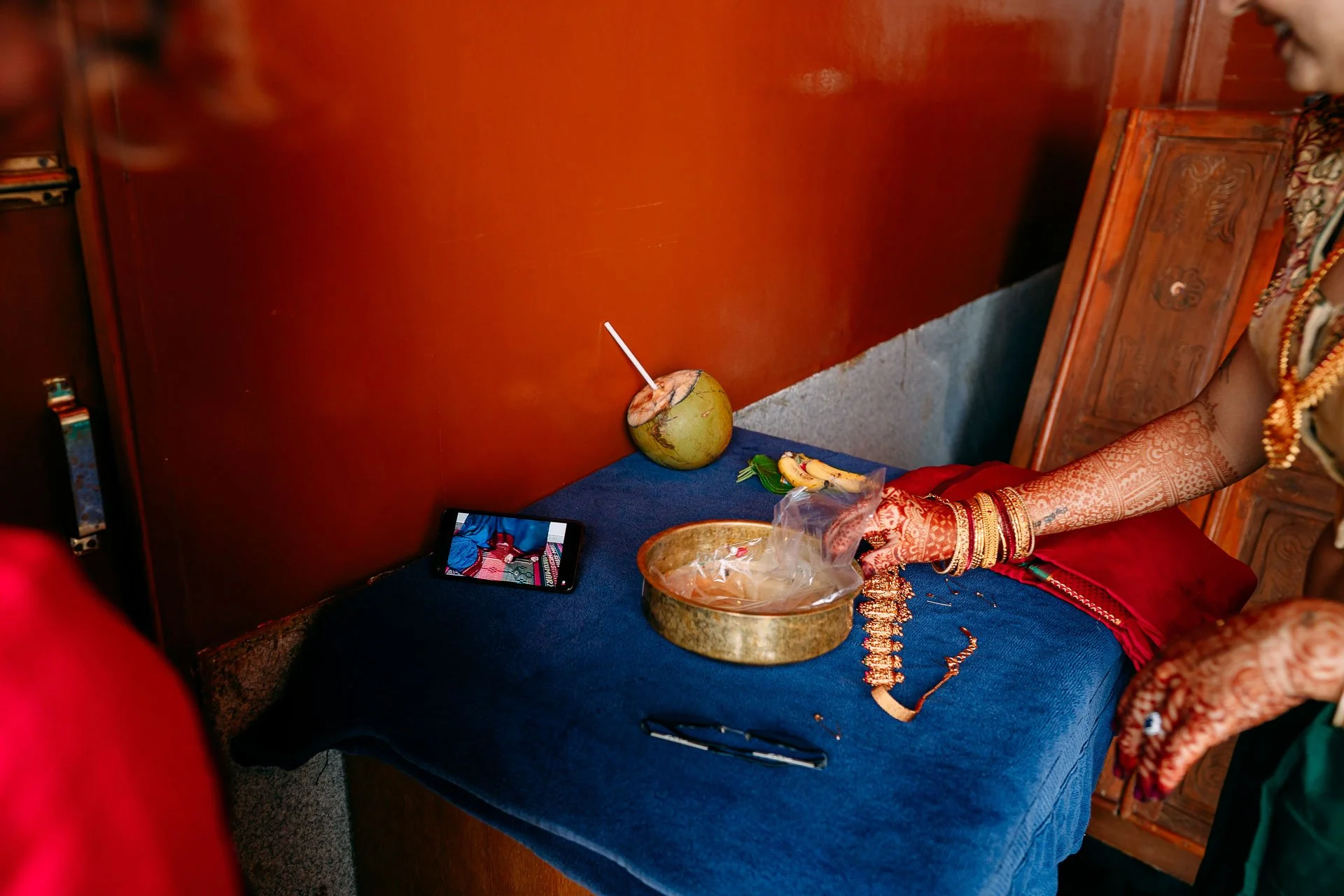 A woman dressed in traditional attire placing offerings on a table covered with a blue cloth, with a coconut adorned with a straw, a smartphone, and various items, including a plate of bananas, on the table.