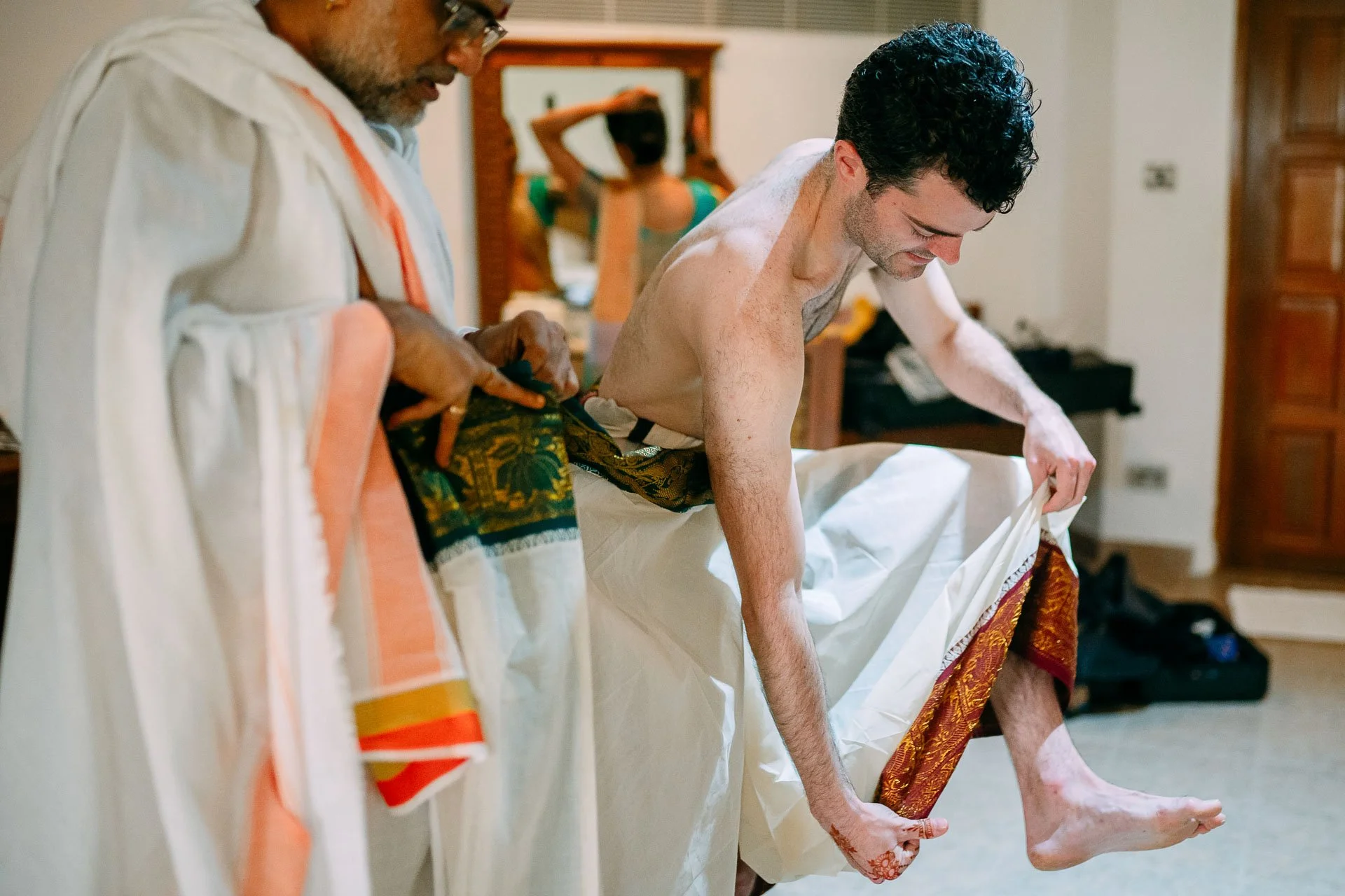A shirtless man with black curly hair dressed in traditional Indian attire is helping to lift his wedding sari while seated. A woman in a green dress is visible in the background, looking into a mirror and adjusting her hair. The setting is a warmly 