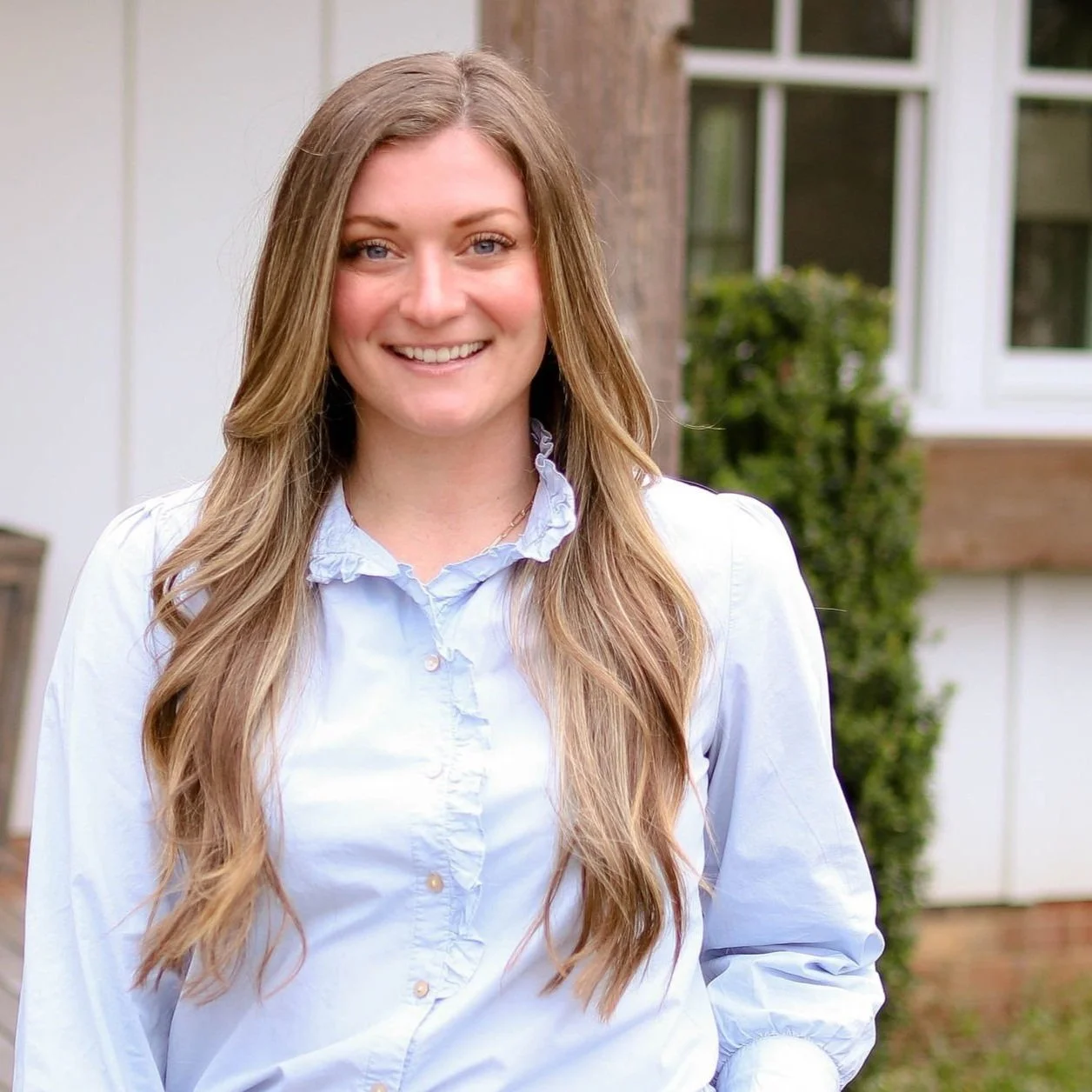 Woman with long, light brown hair and a white blouse standing outside in front of a house with white walls and windows.