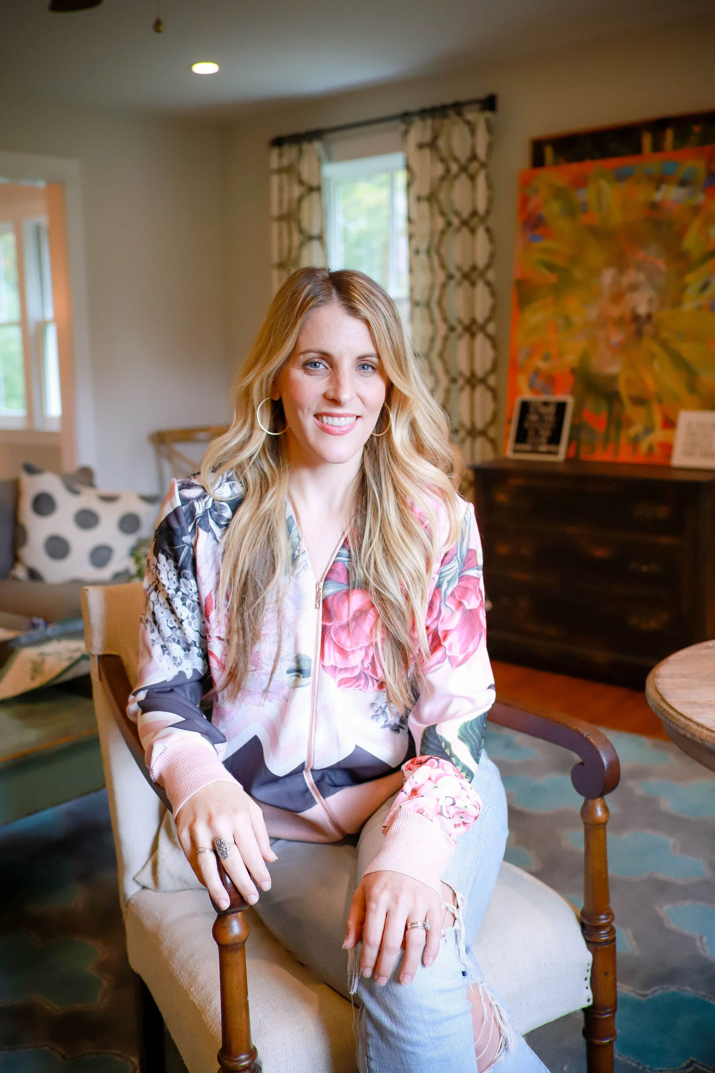 A woman with long blond hair, wearing a floral bomber jacket and ripped jeans, sitting on a beige chair in a well-lit room with patterned curtains, colorful artwork, and a polka dot pillow in the background.